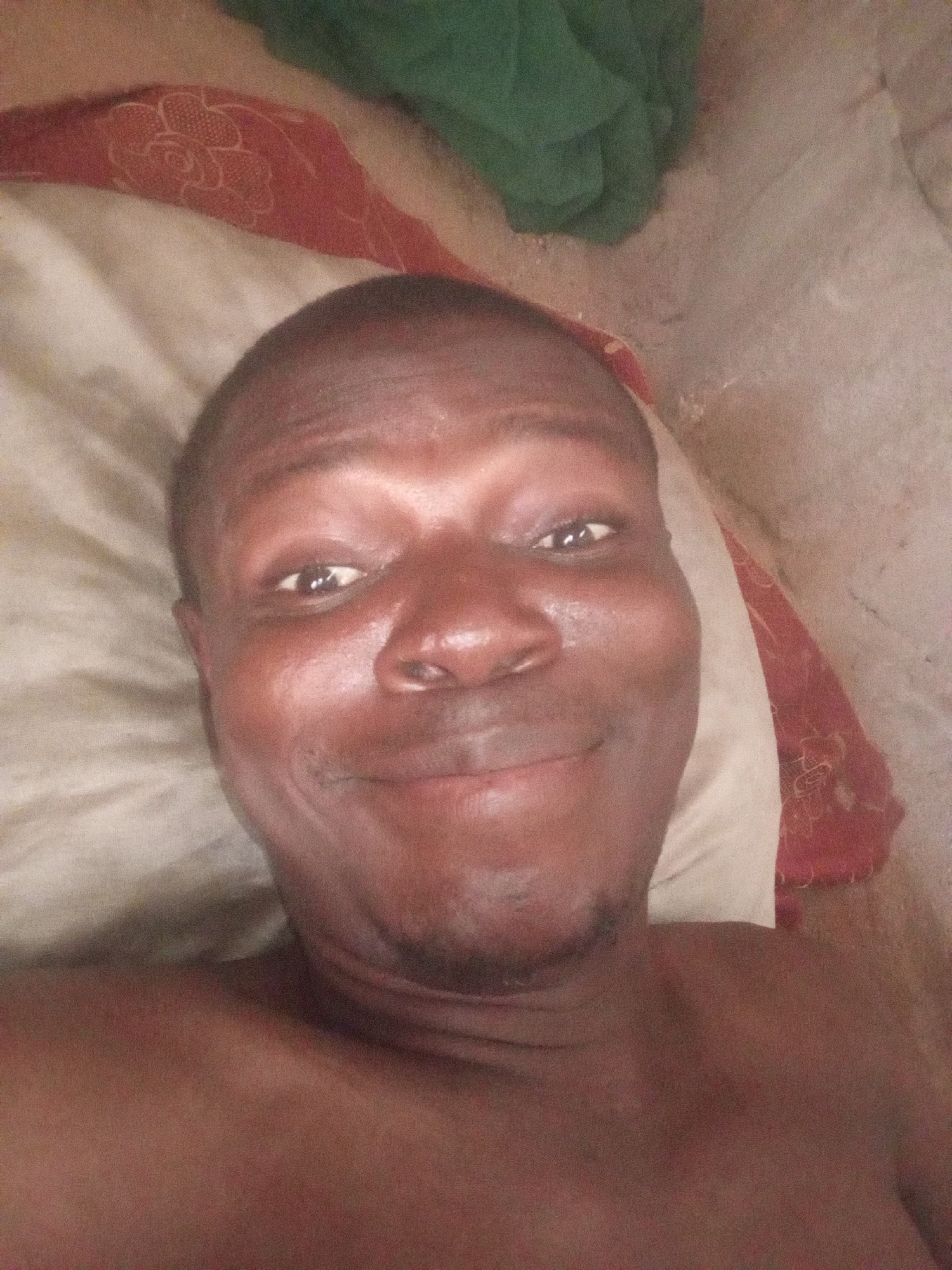 A medium shot depicts an adult man lying on a pillow. His skin is brown, and his head is bald. He is looking directly at the camera with a subtle smile. A green cloth is visible in the upper left corner of the frame, while the pillow beneath him has a reddish patterned design. The location appears to be a bedroom or sleeping area in an unknown city.