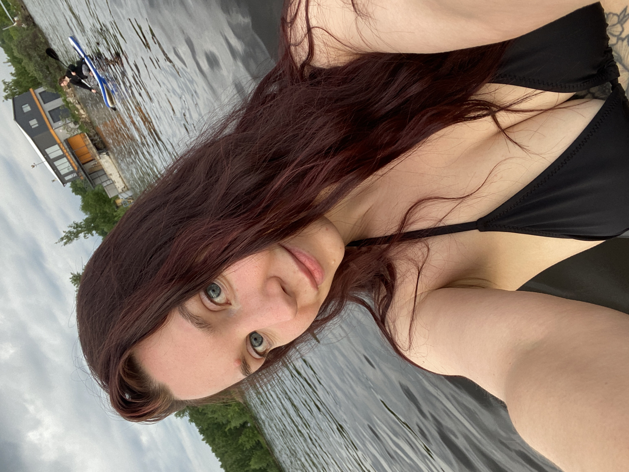 An outdoor, rotated selfie image captures a woman in the foreground and a water scene in the background. The woman, with long, wavy dark red hair and blue-green eyes, looks directly at the camera with a slight smile. She is wearing a black bikini top, with her hair appearing damp, suggesting recent water activity.
Behind her, on a calm body of water, likely a lake or river in Chaikovsky, Russia, another individual is paddling a blue and white kayak or canoe. On the distant shore, a modern, multi-level building with dark siding and large windows stands amidst green trees. The sky is overcast with grey clouds, indicating a cloudy daytime setting. The image portrays a relaxed, leisure activity near water, possibly during summer or warm weather. No discernible text is visible in the image.