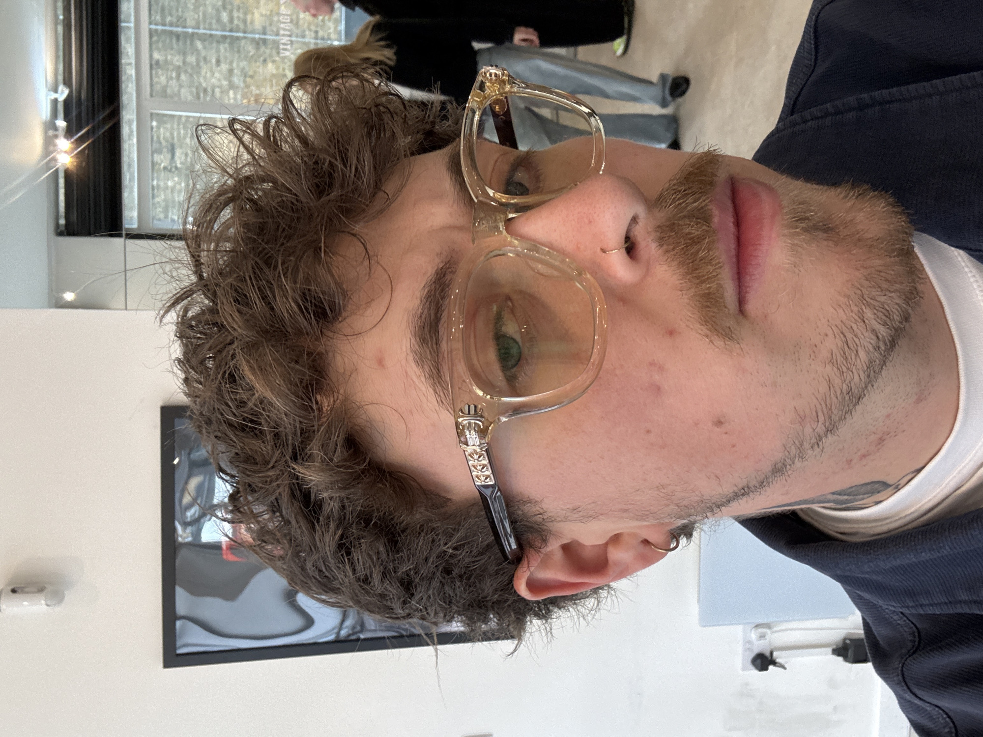 A close-up portrait captures a young adult male with curly, light brown hair and a light brown beard and mustache, looking directly towards the viewer. He wears clear, light amber-tinted eyeglasses with ornate, possibly metallic, temple arms featuring a distinct design, potentially a cross motif. His left eye, which appears green, is clearly visible through the lens. He has a small gold hoop nose ring in his left nostril and a matching earring in his left earlobe. His expression is neutral.
He is dressed in a dark blue jacket or top worn over a white undershirt. The photograph appears to be taken indoors, in a brightly lit setting, possibly a retail space or studio in London, United Kingdom. The background is mostly out of focus, revealing light-colored walls and a large window. Through the window, blurred outdoor elements are visible, and the word "VINTAGE" can be seen reflected in white text on the glass, suggesting the presence of a vintage store or a similar establishment.
Bright reflections are present on various surfaces in the background, including the window, indicating strong natural light, likely during daytime. A dark-framed object, possibly a picture or screen, is partially visible on the wall behind him. The man's hair is somewhat disheveled, and his skin shows a few minor blemishes. The image is captured at a slight upward angle.