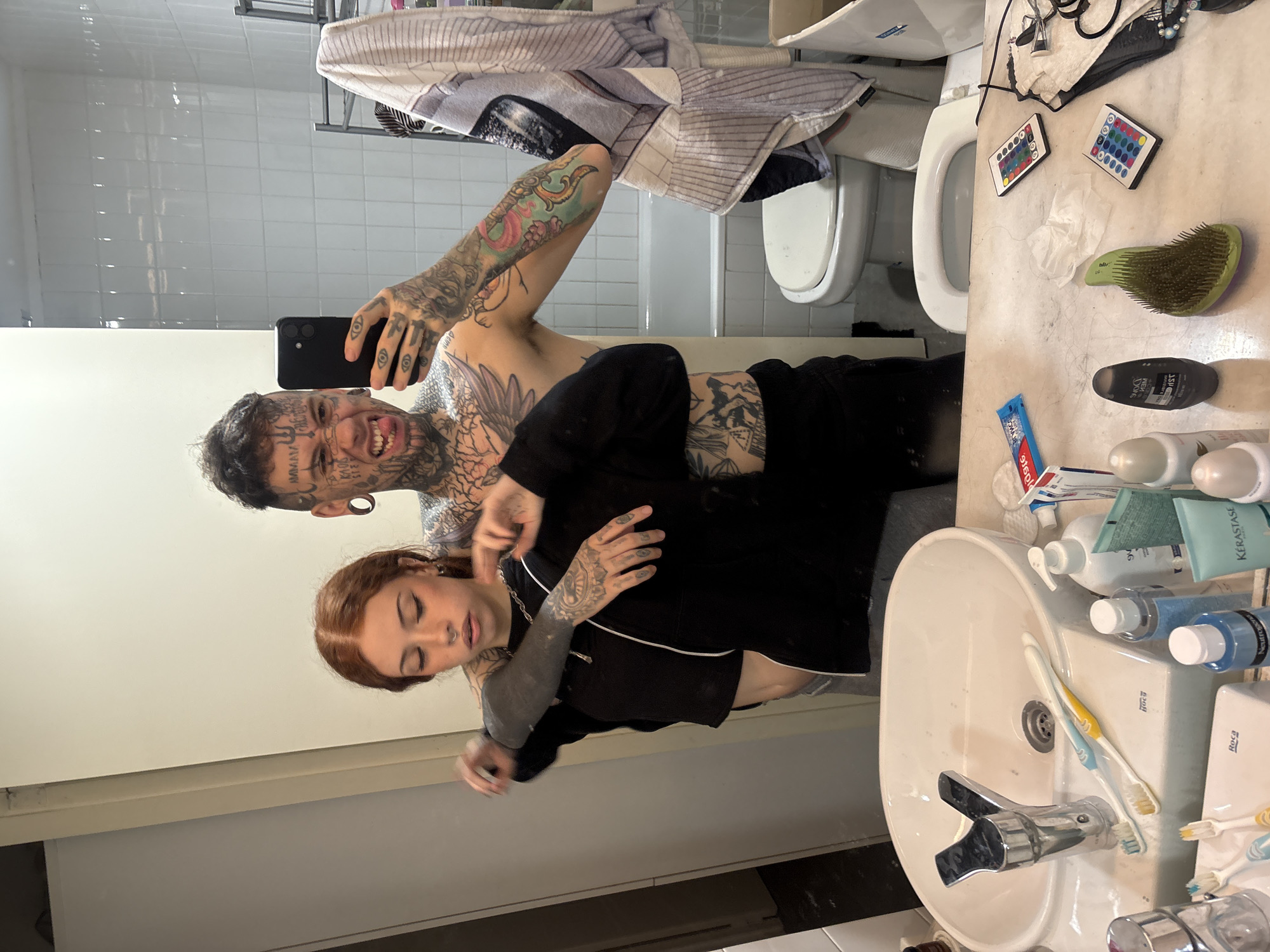 The scene depicts two individuals captured in a mirror selfie within a residential bathroom in the city of Buenos Aires, Argentina.
The person on the right, a male, is heavily tattooed across his face, neck, chest, and arms, featuring numerous black ink designs, including text, symbols, and intricate patterns. He has dark, short hair, prominent ear gauges, and is making a playful or aggressive facial expression with his tongue partially extended and teeth bared. He is shirtless, revealing extensive body art, and appears to be wearing dark shorts or trousers. He holds a black smartphone in his right hand, pointed towards the mirror to take the photograph.
The person on the left, a female, has reddish-brown hair and a nose ring. She has tattoos on her arms, including a solid black sleeve on her left arm and a detailed blackwork pattern on her right forearm. She wears a black long-sleeved top with a white zipper. Her eyes are partially closed, and her expression is subdued. The male has his left arm draped over her shoulders, embracing her from behind, while his right arm, holding the phone, also appears to be around her. Her arms are raised, with her hands near her chest, as if adjusting her clothing or resting.
The immediate foreground displays a white ceramic sink, branded "Roca," with a chrome faucet. Two toothbrushes, one blue and white, and one yellow and white, rest on the sink's edge. The beige-marbled countertop around the sink is cluttered with various toiletries, including bottles of shampoo, conditioner, lotions, toothpaste (Colgate brand visible), cotton pads, and a green-bristled hairbrush. Two small remote controls with colorful buttons, likely for LED lights, and a crumpled tissue are also on the counter to the right.
The background shows a bathroom interior. White subway tiles cover the wall behind the individuals. To the right, a white toilet with its seat down is visible. Behind the toilet, a white bathtub is present. A metal towel rack holds a striped, multi-colored towel. The floor is visible in parts, appearing as dark grey or black tiling. The reflection in the mirror further reveals portions of the bathroom, including another toilet bowl below the main one, indicating a clean and functional, albeit somewhat lived-in, domestic setting.