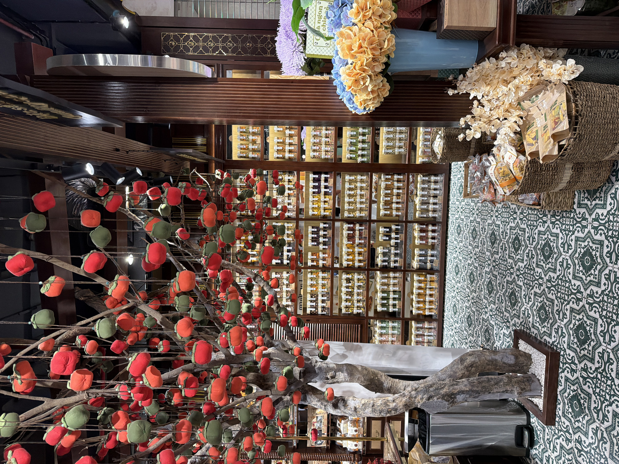 A street-level interior scene in Da Nang, Vietnam, features a decorative tree adorned with red and green fabric ornaments that resemble fruit, hanging from branches and the ceiling. The tree is placed in a wooden planter filled with white pebbles. Behind the tree, shelves are stocked with numerous glass jars containing various food items, likely spices or dried goods. To the right, a display of flowers includes light blue and orange hydrangeas in a blue vase, and purple flowers. Woven baskets filled with packaged food items are also visible on the floor. The floor is covered in a patterned tile with a green and white design. The overall impression is that of a market stall or a shop specializing in food products, possibly in a tourist-friendly area of the city. There are no people visible in the frame.