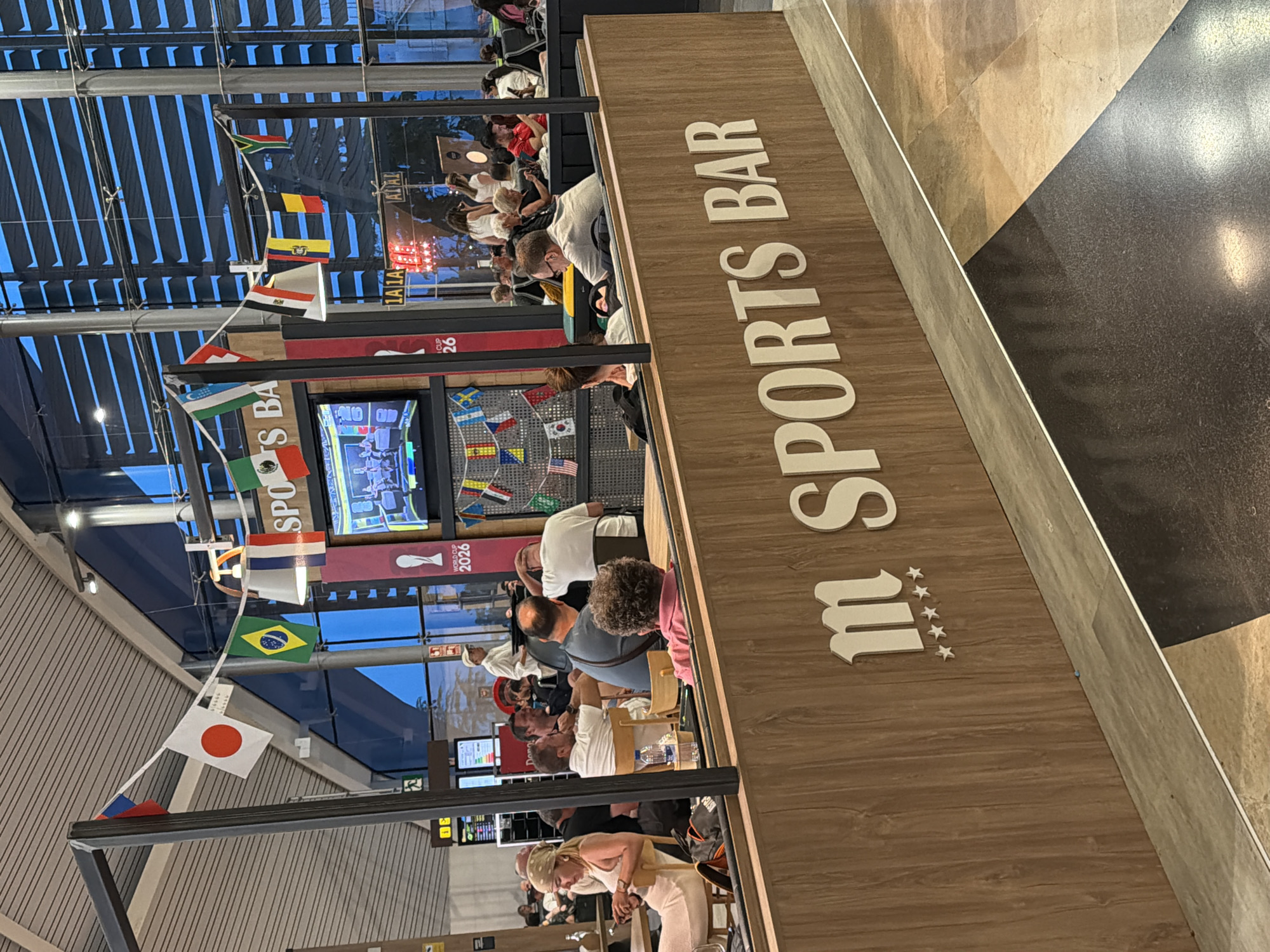 The image depicts an indoor scene at a sports bar. A wooden counter with the words "m SPORTS BAR" and five star symbols is prominently featured in the foreground. Behind the counter, several people are seated at tables and chairs, some of whom are engaged in conversation or looking at a screen. A television displaying what appears to be a sports broadcast is visible in the background, along with hanging flags of various countries. The environment suggests a public space, possibly an airport or a large entertainment venue, with modern architecture and lighting. The overall atmosphere appears casual and social.