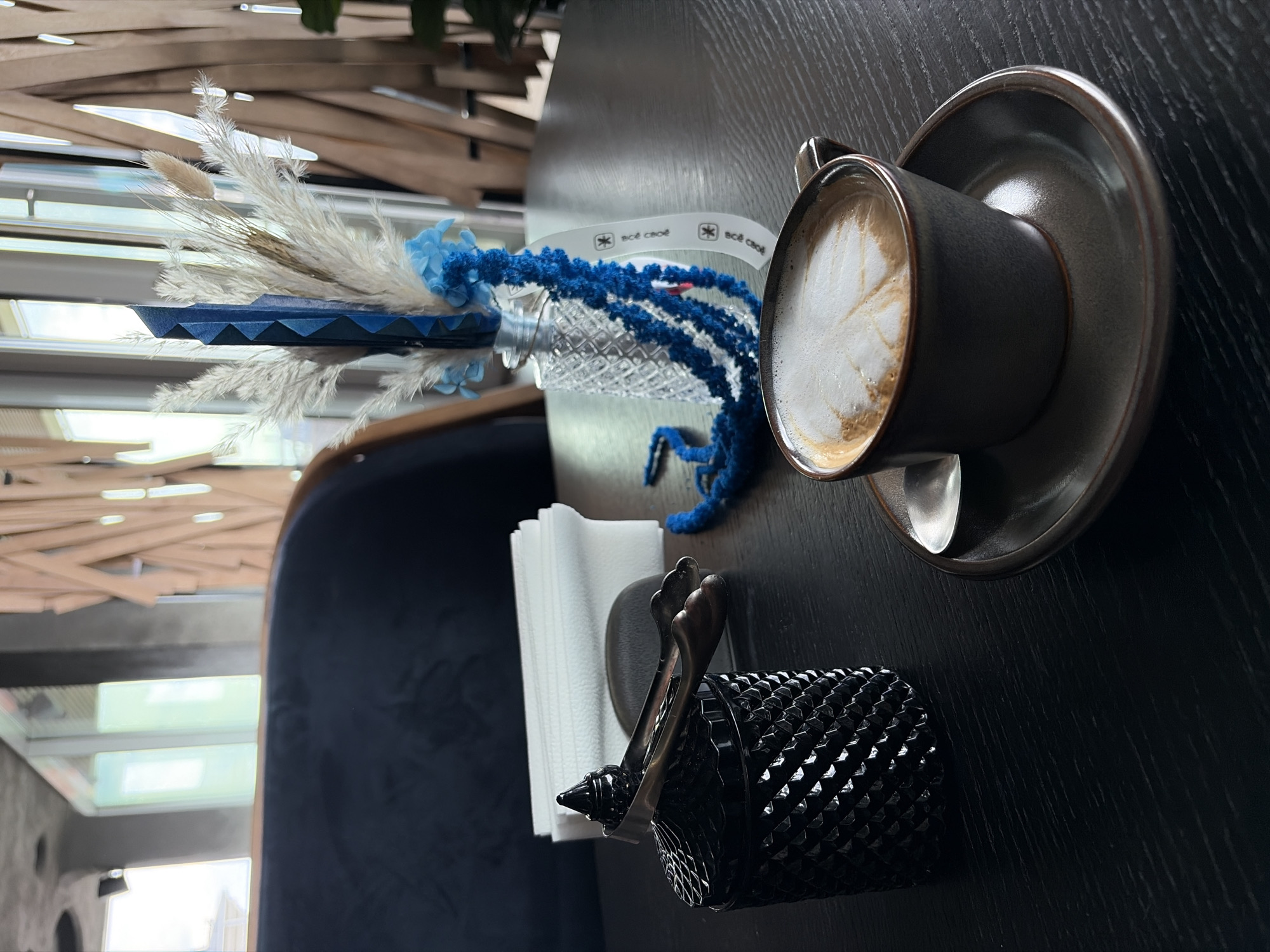 The image displays an indoor table setting, likely within a cafe or restaurant, viewed from a slightly elevated and angled perspective. The primary surface is a dark, possibly wooden, tabletop.
On the right side of the table, a dark grey ceramic coffee cup with a matching saucer holds a frothy beverage, likely a latte or cappuccino, featuring delicate latte art. A silver spoon rests within the saucer.
Towards the center, a decorative arrangement sits on the table. It includes a small, blue, trapezoidal vase holding white and light brown dried grasses or pampas grass, complemented by small blue artificial flowers. Next to this, a clear, textured, possibly glass or plastic, object supports a length of fuzzy blue yarn or string that drapes over it and onto the table.
In the foreground on the left, a stack of white paper napkins is held in a distinctive black holder with a faceted or diamond-patterned texture. A pair of small silver tongs rests on this holder.
In the background, blurred architectural details are visible, including wooden slats or beams, suggesting a ceiling or wall design, and reflections of light through a window or opening. A dark blue upholstered chair or booth is partially seen on the far left.
A white ribbon, positioned behind the blue yarn, clearly displays the repeated Russian text "всё свое" (vse svoe), which translates to "all mine" or "everything is mine," accompanied by a star-like logo. The scene conveys a calm, casual atmosphere, typical of a daytime coffee break. The location context for this media is Izhevsk, Russia.