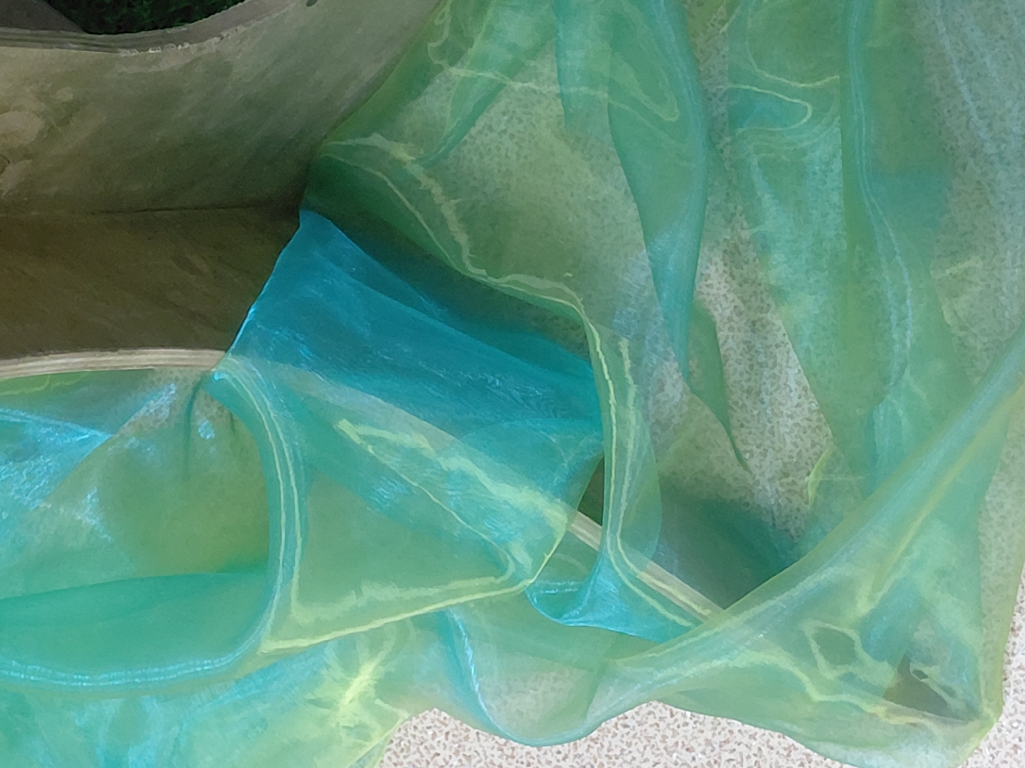 This close-up image presents an abstract composition dominated by a sheer, iridescent fabric, likely organza or a similar synthetic material. The fabric exhibits a striking gradient of colors, transitioning from vibrant turquoise and emerald green to lighter lime green and yellowish-green hues, suggesting a shifting color palette depending on light and layering.
The fabric is intricately draped and folded, creating numerous overlapping layers and soft curves. These folds and wrinkles enhance the translucent quality of the material, allowing varying degrees of light to pass through and reflect, resulting in areas of intense color saturation and other areas where the underlying surfaces are subtly visible. The interplay of light on the glossy surface creates bright, almost white highlights along the edges of the folds.
The background consists of two main elements: a lighter, textured surface, possibly a floor or tabletop with a fine, granular pattern, and a darker, more indistinct area in the upper left, which appears to be a shaded space or an interior wall. No specific objects or identifiable settings beyond the fabric itself are discernible.
There is no activity or event taking place, and no people are present. The image captures the static beauty and textural richness of the material. No text is visible in the media file. The lighting appears soft and diffused, highlighting the fabric's luminous qualities without casting harsh shadows, making it difficult to determine the specific time of day or weather conditions. There are no direct location cues within the image itself.