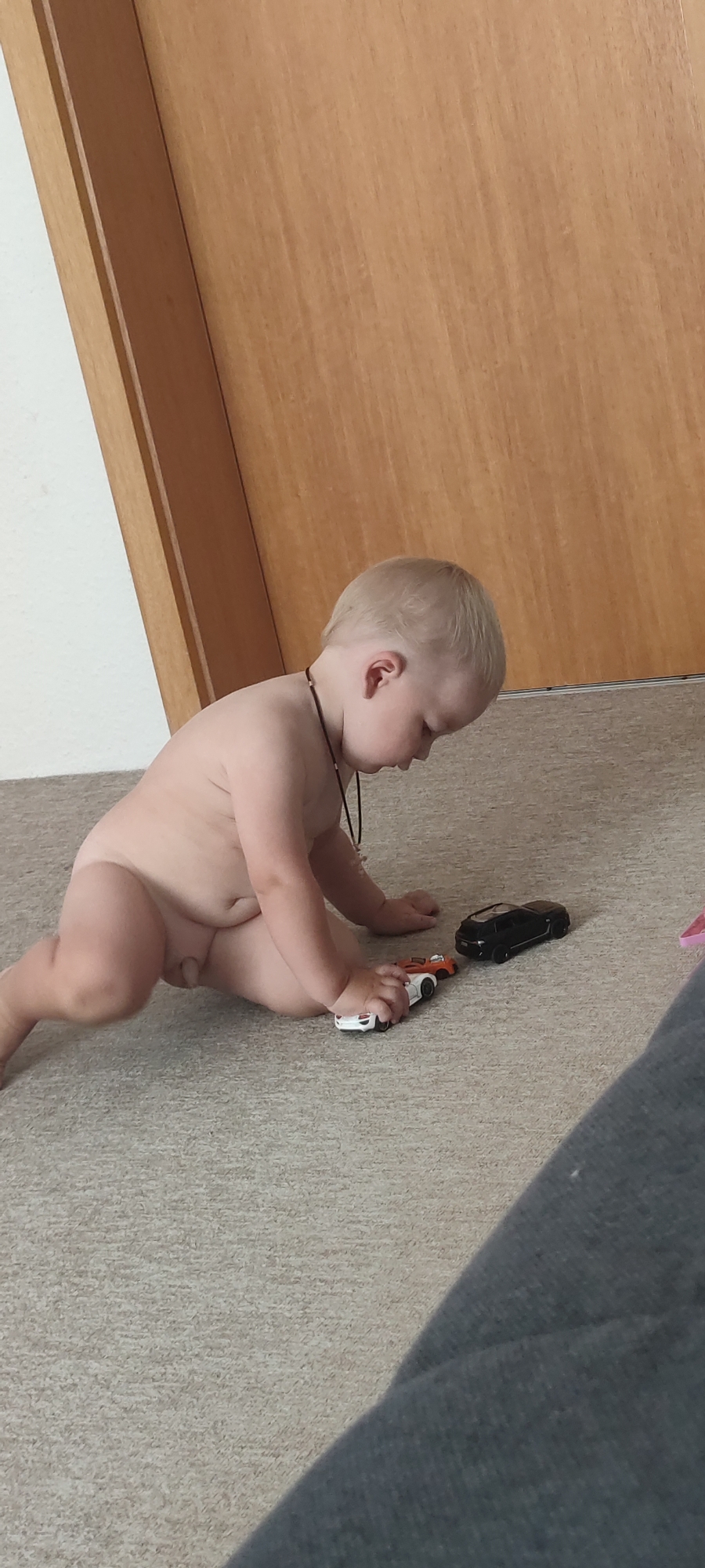 A naked baby is playing with toy cars on a carpeted floor. The baby has blonde hair and is leaning forward, looking down at the toys. There are three toy cars visible: a white and orange one, a white one, and a black one. The setting appears to be indoors, with a wooden door in the background. There are no other people visible in the image. The lighting suggests it might be daytime. No text is visible. The baby appears to be focused on the toys. The location is Liebenburg, Germany.