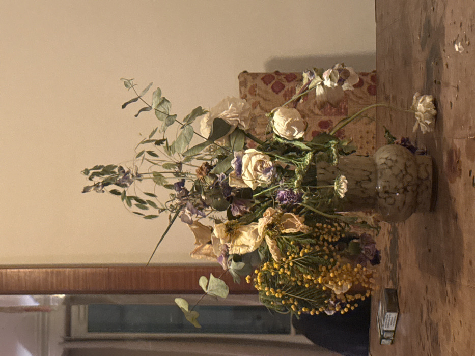 The image displays an indoor still life scene, presumably located in Paris, France. A large arrangement of dried and decaying flowers, including wilted white and cream roses, yellow mimosa-like clusters, and faded purple or blue blossoms, is presented in a mottled, light-colored ceramic vase with a textured pattern. The vase rests on a dark-stained wooden table exhibiting visible grain and minor surface wear. To the left of the vase, a white and green cigarette pack lies on the table. In the background, a warm, plain cream-colored wall is visible. To the left, a tall, narrow mirror with a wooden frame reflects a blurry interior space, possibly a window or doorway with light-colored curtains. To the right, behind the vase, a section of upholstered furniture or a cushion with a patterned fabric (red/brown floral or geometric motifs on a lighter background) is partially visible. Several dried flower fragments are scattered on the table surface. No individuals are present, and no actions or interactions are depicted. The lighting is soft and ambient.