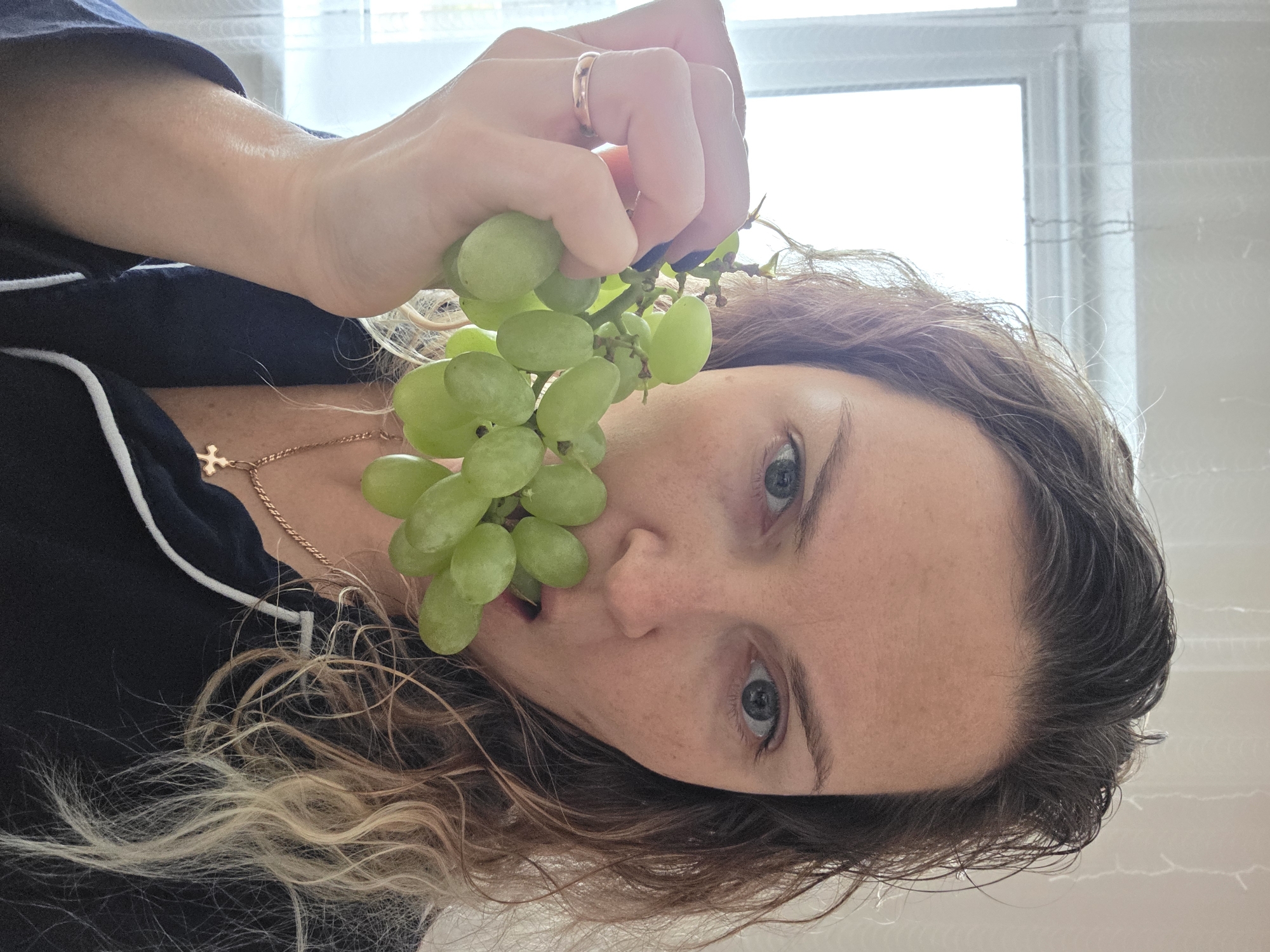 A woman is holding a bunch of green grapes in front of her face. She is wearing a dark blue pajama top with white piping and a gold necklace with a cross pendant. She has a wedding ring on her ring finger. Her hair is brown and blonde. The background is a white wall with a window. There is no evidence to suggest this image was taken in the city of Chaikovsky, Russia.