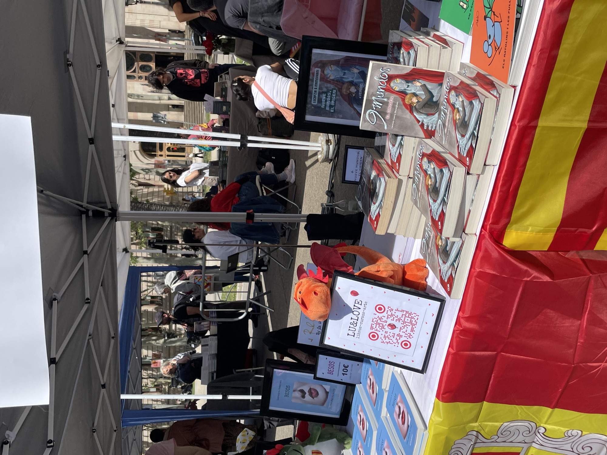 The image depicts an outdoor book fair or market on a sunny day, likely in Barcelona, Spain, given the visible Catalan flag. The foreground is dominated by a vendor's table set up under a white canopy tent.
On the table, stacks of books are prominently displayed. Multiple copies of "NUEVE MUNDOS, EL ORIGEN" by Lucrezia G. Mameghani, featuring a fantasy-style cover, are visible, along with a framed print of its cover. Another book titled "BESOS" by Lucrecia & Rosegton is also present, marked with a price of "10€." Decorative stuffed toys, including an orange dragon-like creature, are arranged on the table. A stand displays a QR code and text reading "LU&LOVE libros-cursos-arte," indicating the vendor's brand. A red and yellow striped flag, clearly identifiable as the flag of Catalonia with part of its coat of arms visible, is draped over the front of the table.
In the midground, several people are seen browsing books or seated under other canopy tents, suggesting a public event. One woman in a white shirt and black leggings with a pink cross-body bag is intently looking at books on a nearby table. The background features architectural details of historic-looking buildings, consistent with an urban European setting. The overall atmosphere appears relaxed and engaging, with attendees exploring the book offerings.