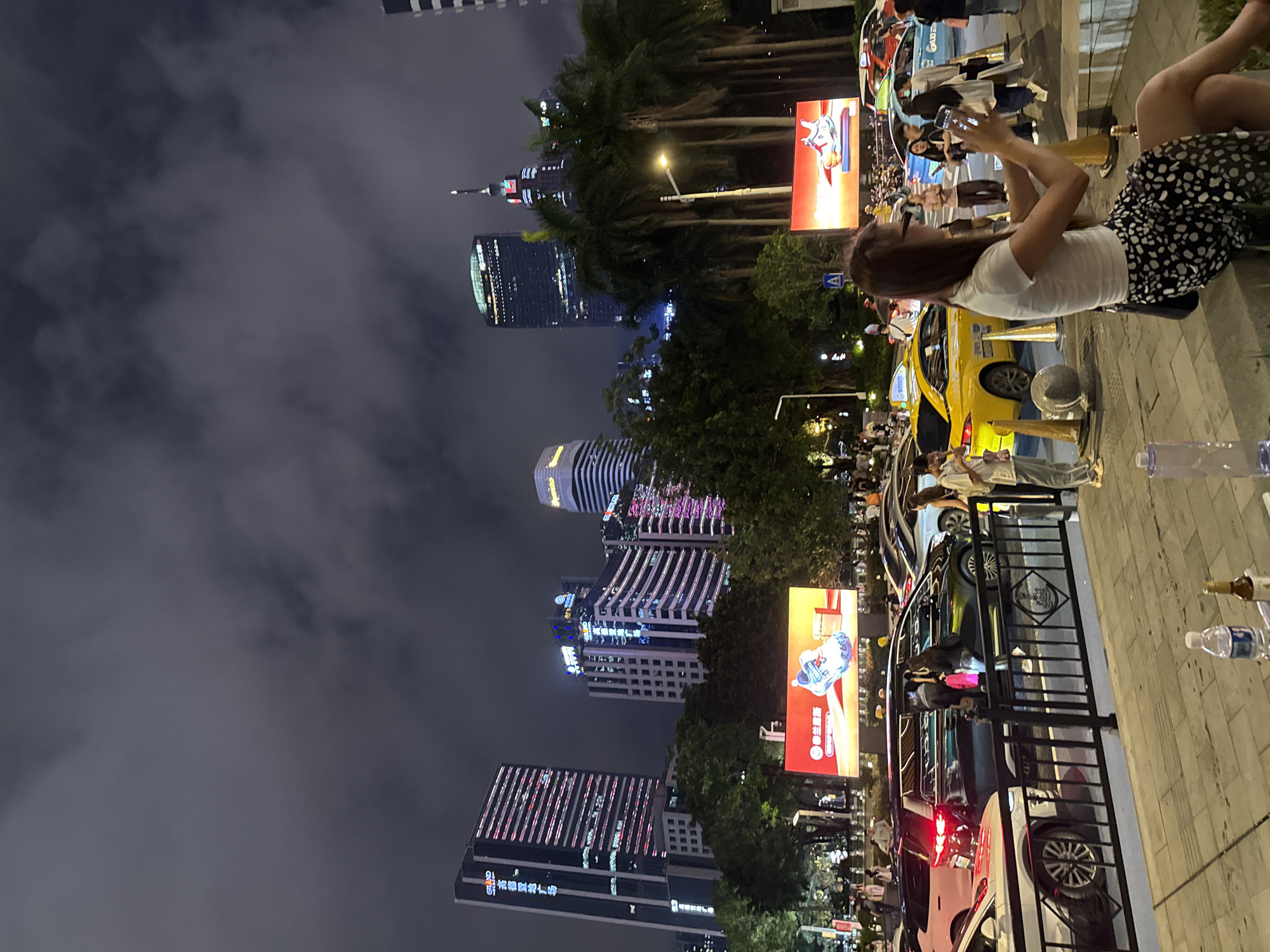 In Tianhe District, China, at dusk, a busy street scene unfolds with a focus on a woman in the foreground wearing a white top and a black and white spotted skirt, sitting on a concrete ledge and holding up her phone, as if taking a picture or recording. She is looking at her phone with her head tilted. Beside her are two empty plastic bottles and a bottle with a dark liquid. Behind her, traffic is congested with cars of various colors, including a prominent yellow taxi and a dark SUV with its doors open. Several people are standing around the SUV, some appearing to be interacting with those inside. In the background, a bright billboard displays an advertisement for shoes. Towering skyscrapers with illuminated windows dominate the skyline, indicating a densely populated urban environment. The sky is dark and cloudy, suggesting nightfall or overcast weather. Palm trees and other foliage are interspersed among the buildings and along the roadside, adding a touch of nature to the urban landscape.