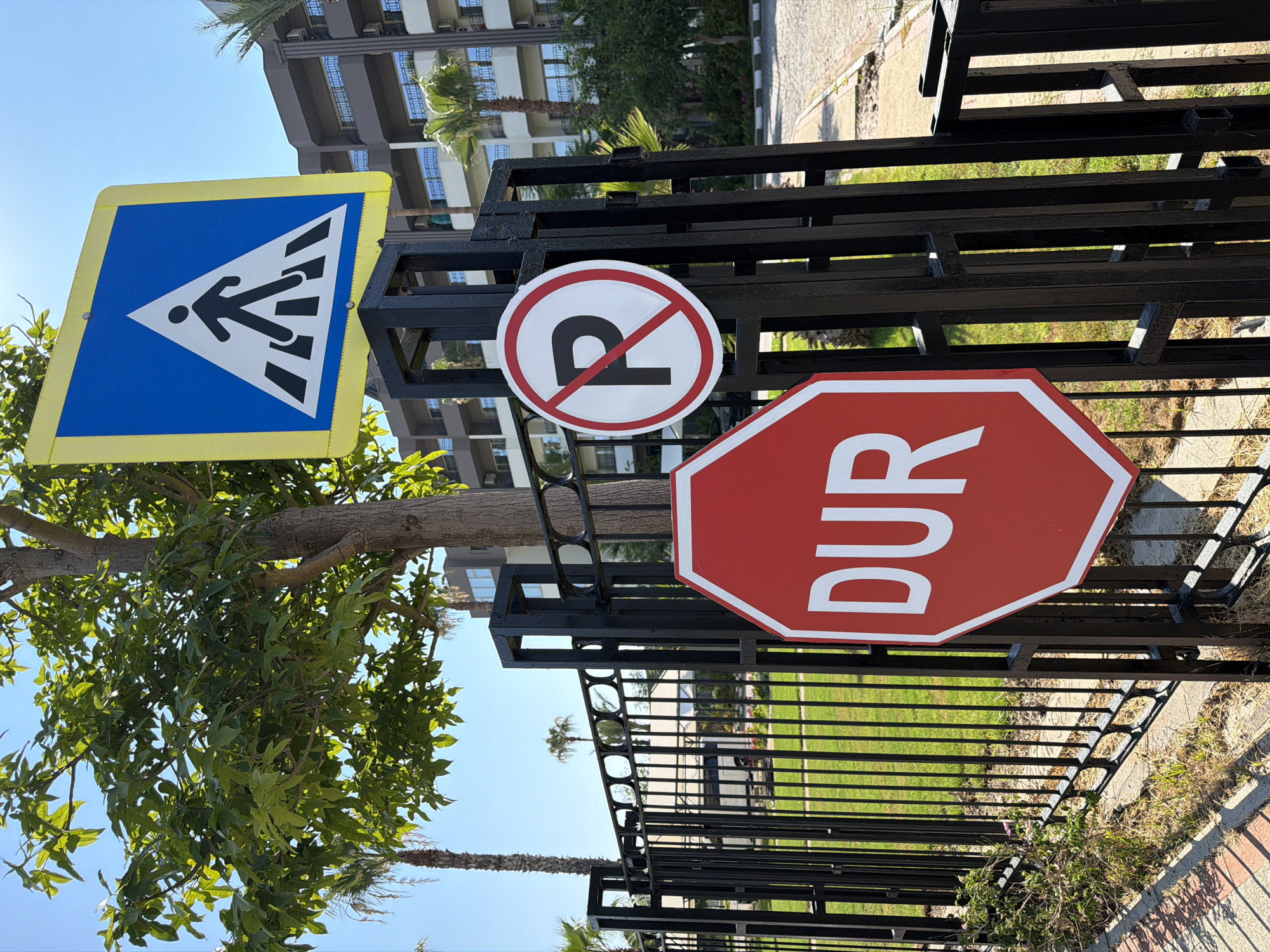 In Kemer, Turkey, a black metal fence is adorned with three traffic signs. At the top, a blue square sign with a yellow border depicts a white pedestrian crossing. Below it, a circular sign with a red border and a diagonal red line across it indicates no parking, with a black letter "P" in the center. A large red octagonal stop sign with the white text "DUR" is positioned at the bottom. A leafy tree with a textured brown trunk stands behind the signs and fence. In the background, a multi-story building with balconies and palm trees are visible under a clear blue sky.