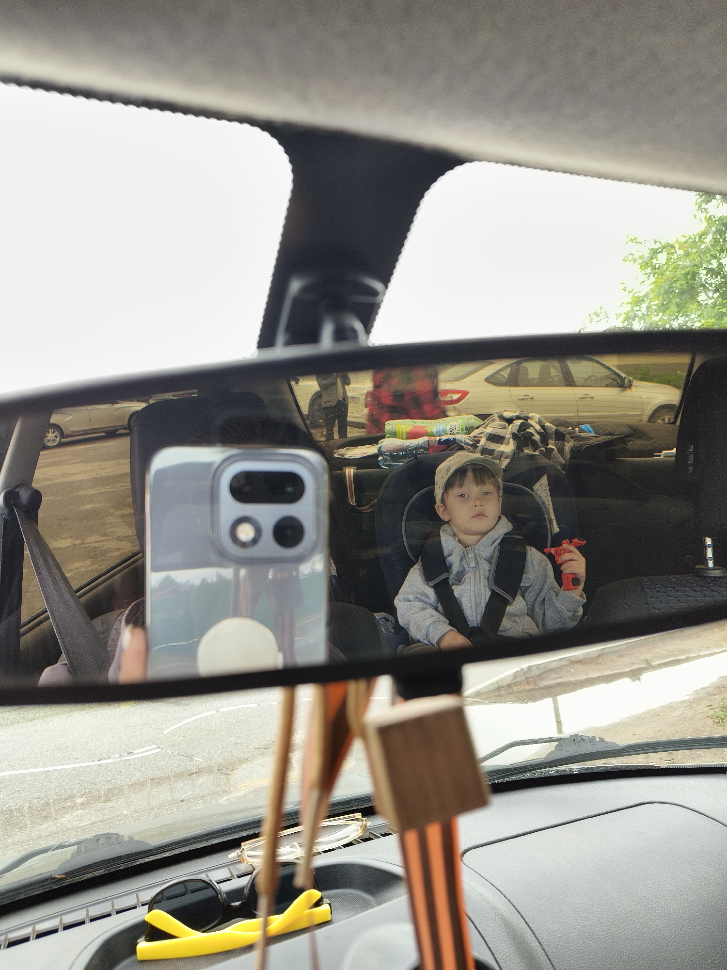 The image shows a view through a car's rearview mirror. Reflected in the mirror is a child seated in a car seat in the back of the vehicle, holding a red toy. There are various items piled behind the child, including what appears to be blankets, water bottles, and possibly other luggage. Also visible in the mirror is the reflection of the exterior of the car, showing what looks like a parking lot, and other parked vehicles. The city's name "Noviy, Russia" is not discernable. In the foreground, the car's dashboard is visible, with sunglasses and other small objects. The rearview mirror is partially obscured by a phone and hanging decorations.