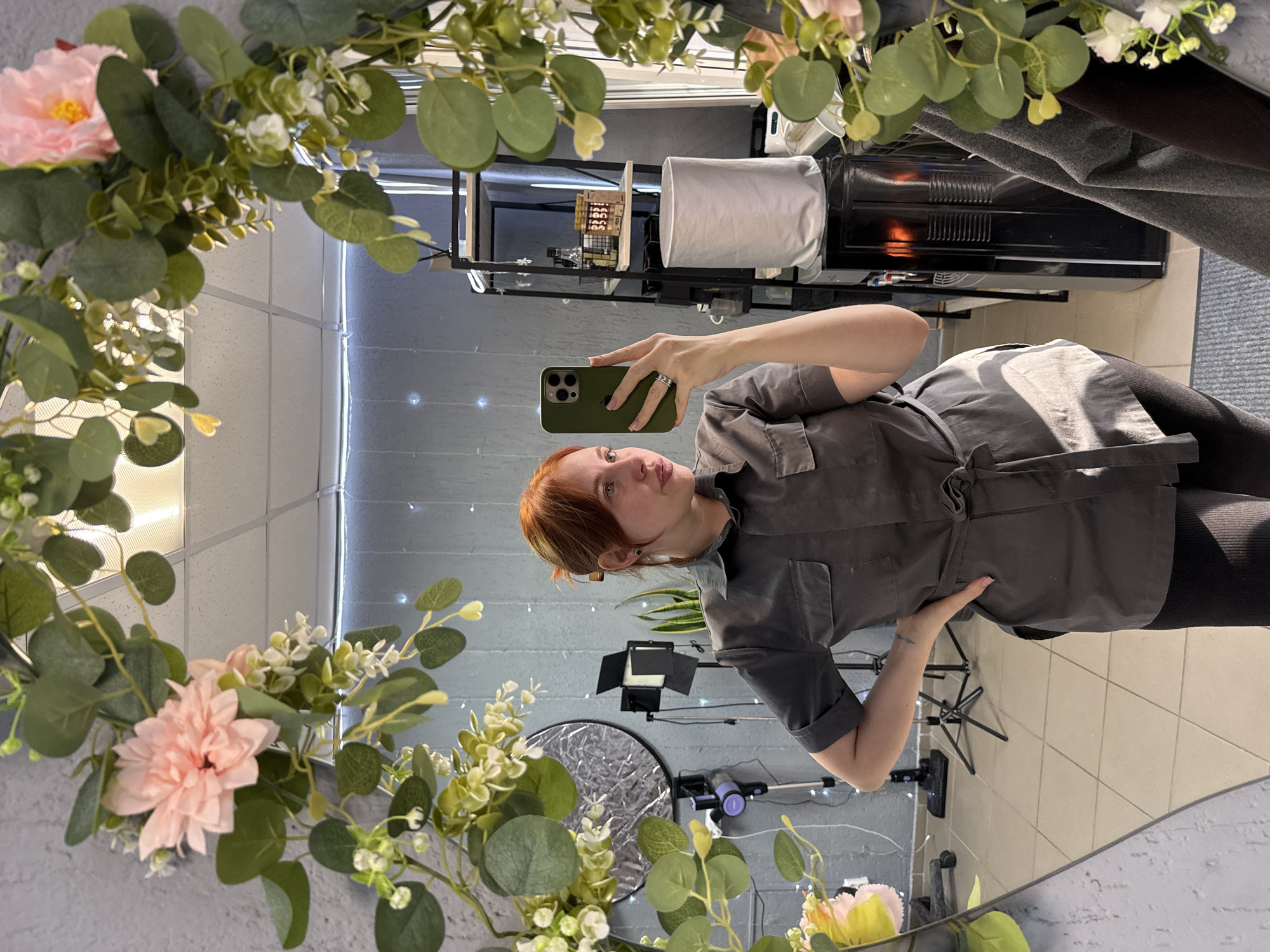 An indoor scene in Chaikovsky, Russia, shows a mirror selfie taken by a female subject. The woman, appearing to be in her 20s or 30s with reddish-brown hair styled back, is wearing a dark grey short-sleeved tunic with breast pockets and a waist tie, over black fitted pants. She holds a green-cased smartphone in her left hand, angled to capture her reflection, while her right hand rests on her hip. A dark stud earring is visible in her left ear, a ring on her left ring finger, and a tattoo on the back of her right hand.
The irregular-shaped mirror is decorated with artificial green foliage, pink flowers, and white floral accents. The reflected environment reveals a textured grey wall on the left, displaying a silver circular photography reflector on a stand, a black studio light with barn doors on a stand, a purple stick vacuum cleaner, a potted snake plant, and horizontal string lights. On the right, a black metal shelving unit holds various items, including bottles, jars, a basket, and a small digital clock. Below the shelving, a white cylindrical cover sits atop a black water cooler, which exhibits an orange internal glow. A partial view of a window with vertical blinds and dark garments hanging on a rack are also visible. The floor consists of light-colored square tiles, with a blue-grey mat in the foreground. Overhead, the ceiling features white suspended panels with visible fluorescent light fixtures.