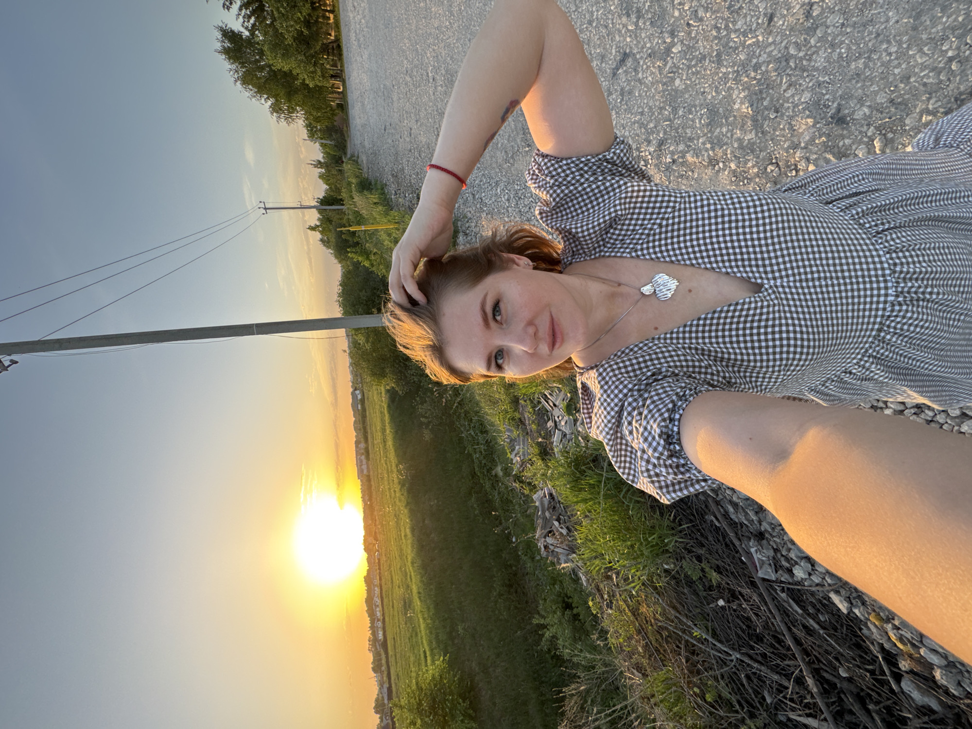 A woman is depicted in an outdoor setting with a visible sunset, possibly in or near Горный, Russia. She is wearing a checkered dress and a necklace with two silver pendants. She is lying on a surface that appears to be a combination of gravel and vegetation. Her right hand is raised to her head and has a red bracelet on her wrist. In the background, there is a field, trees, and utility poles silhouetted by the sunset.