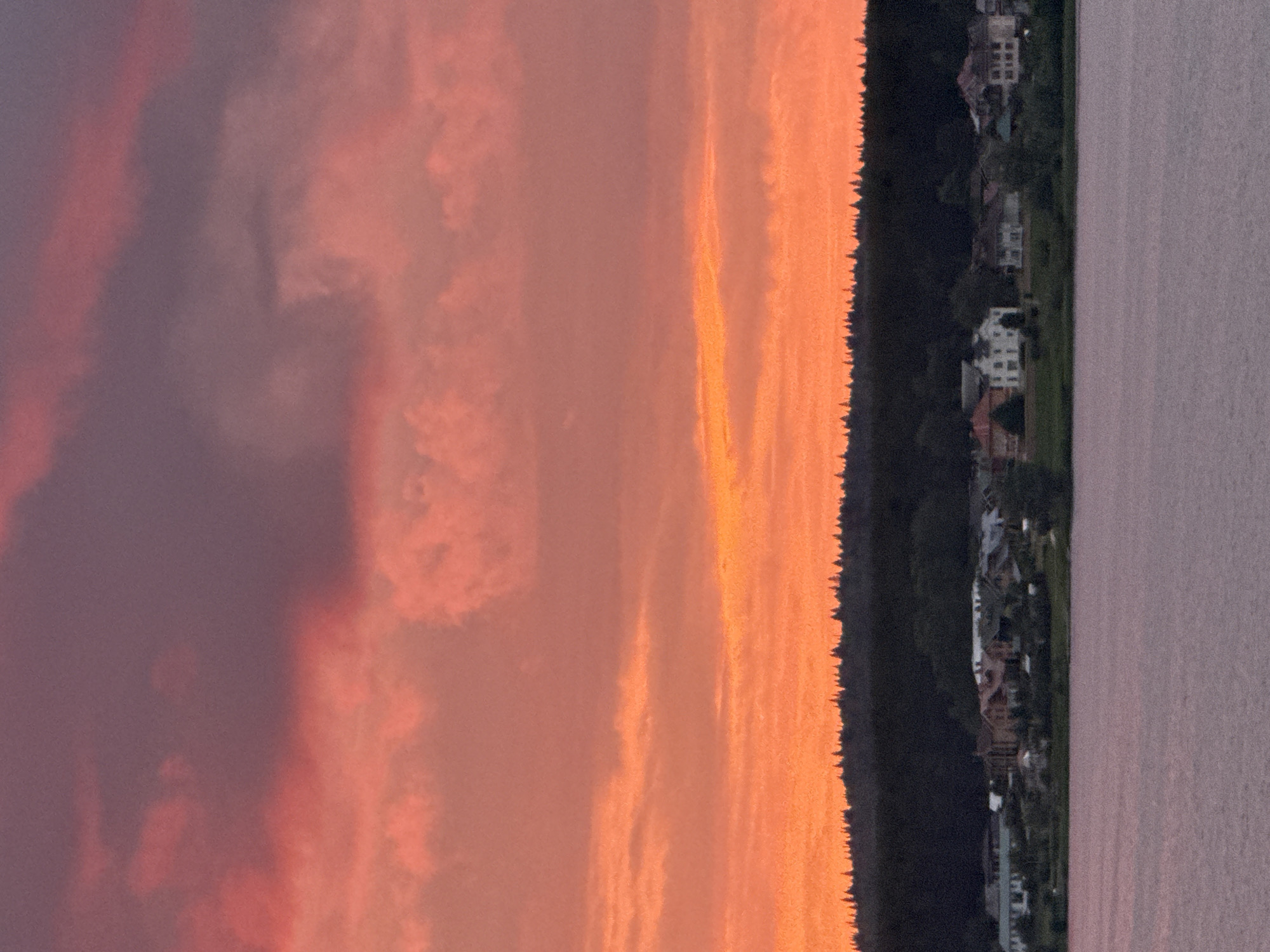 The image shows a picturesque sunset over the city of Votkinsk, Russia. The sky is ablaze with vibrant hues of orange, pink, and purple, casting a warm glow over the landscape. In the foreground, a serene body of water reflects the colorful sky. Beyond the water, a lush green forest stretches along the horizon, dotted with the silhouettes of numerous coniferous trees. Nestled at the edge of the forest is a charming village, characterized by its array of houses with red and white roofs, nestled among trees and greenery. The overall scene is tranquil and idyllic, capturing the beauty of a Russian sunset.