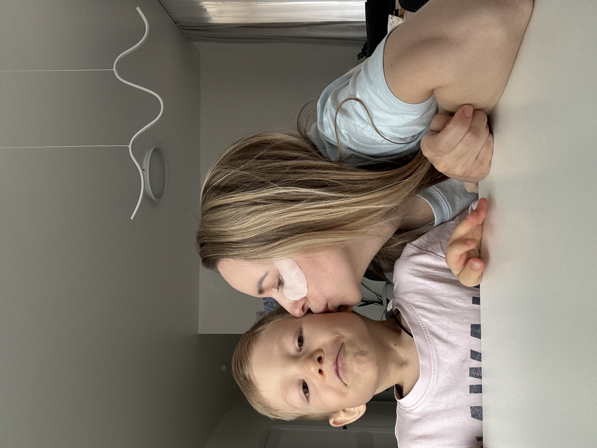 The image shows a woman kissing a young child on the cheek. The woman has long blonde hair and is wearing a light blue shirt and an eye mask. The child has blonde hair and is wearing a light pink shirt with dark stripes. The background includes a white table, a gray wall, and a white modern-style light fixture. While it might appear to be set in an interior space in the city of Chaikovsky, Russia, there is insufficient visual information in the image to verify or confirm this location.