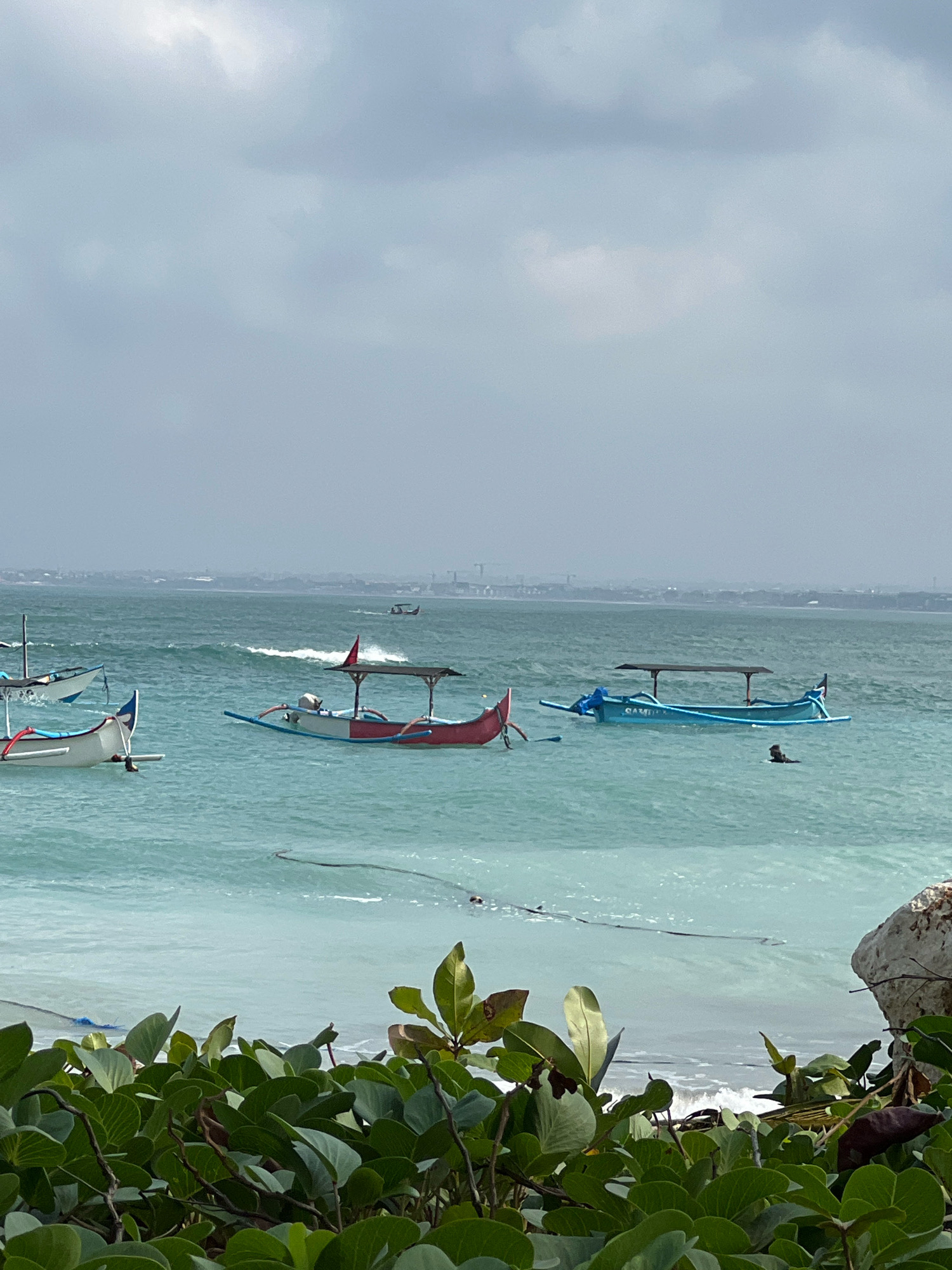 A photograph taken from a slightly elevated position shows the coastline of Kuta, Indonesia. In the foreground, lush green foliage with large leaves obscures the bottom of the frame. Beyond the vegetation, the turquoise ocean stretches out, with gentle waves breaking near the shore. Several traditional outrigger boats are visible on the water. One white and blue boat is positioned on the left, with its mast visible. Closer to the center, a red and white boat with a dark canopy is floating. To the right, a light blue boat with a dark canopy has the word "SAMUDRA" written on its side. A person is swimming in the water near this blue boat. In the background, a hazy cityscape with buildings and structures can be seen along the horizon, under a cloudy sky.