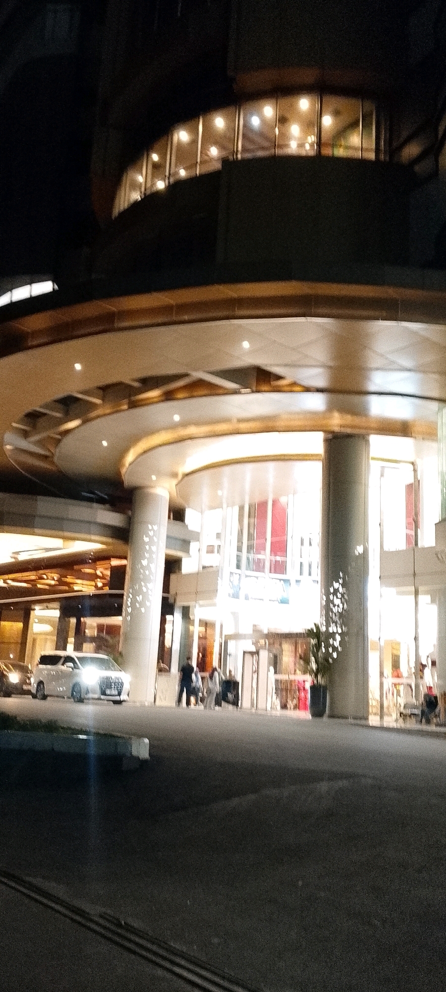 The image captures a nighttime view of a modern building entrance, likely a hotel or commercial complex, located in Depok, Indonesia. The architecture features a curved, multi-tiered facade with extensive glass windows and prominent circular columns. Overhead, curved awnings illuminated by warm, recessed lighting create a layered effect. In the foreground, a white SUV with its headlights on is parked or slowly driving on the asphalt. Several people are visible near the building's entrance, some appearing to be walking with luggage, suggesting travel or arrival. The overall scene is well-lit by artificial lights, creating a reflective quality on the polished surfaces of the building and the road.