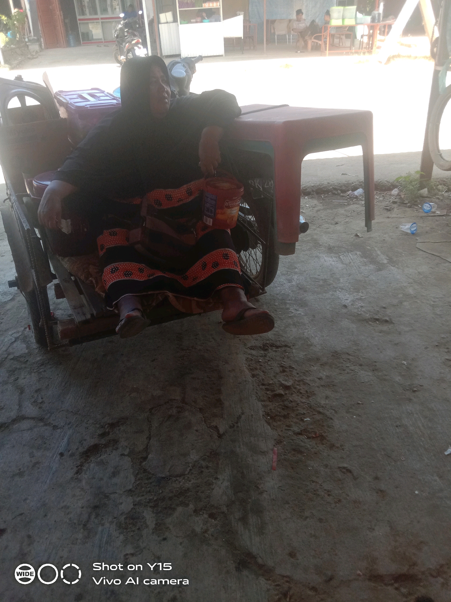 An elderly Indonesian woman, wearing a black hijab and a patterned skirt, is seated in a pedicab. The pedicab appears to be stationary on a paved surface, possibly a street or courtyard, in front of a building with open stalls. The woman is leaning back, with her left arm resting on the edge of the pedicab. A plastic container and a bottle are visible within the pedicab. In the background, other people and various objects including chairs, tables, and a motorcycle are present. The environment suggests a warm, outdoor setting. The image is watermarked with "WIDE Shot on Y15 Vivo AI camera."