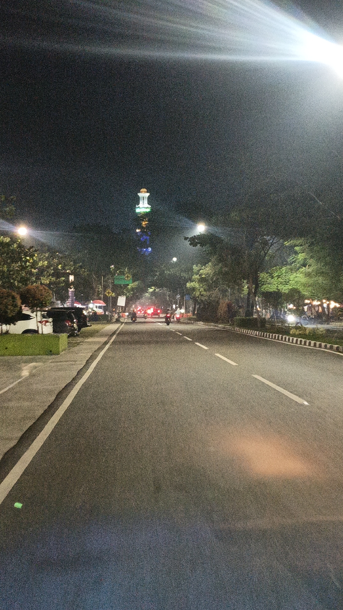 The image captures a nighttime street scene in Sodong, Indonesia, dominated by a wide asphalt road. A tall, illuminated tower, possibly a landmark or monument, stands prominently in the distance, bathed in green and blue lights. The road is lined with trees on both sides, their foliage illuminated by streetlights and the distant tower. Vehicles, primarily motorcycles, are visible on the road, their headlights and taillights creating streaks of light. Some cars are parked along the left side of the road. The overall atmosphere is one of a well-lit urban street at night, with the tower serving as a focal point.