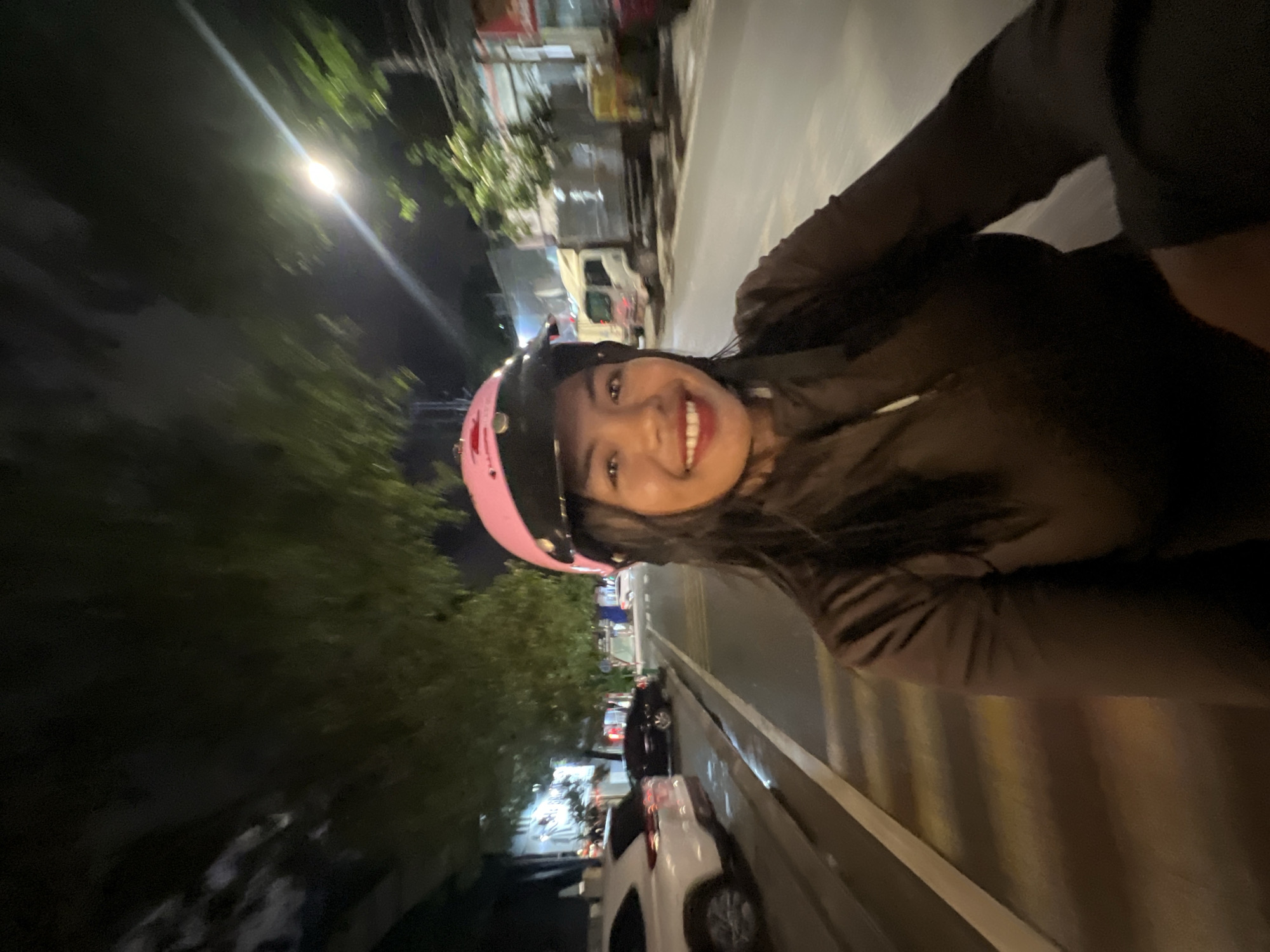 A woman is on a motorbike at night, wearing a pink helmet and a dark brown jacket. She is smiling and looking at the camera. The street is wet, suggesting it has rained recently. There are trees, parked cars, and a truck visible in the background. The scene appears to be in a city. Based on visual cues such as vehicle types and road markings, this scene is consistent with a location in Minh Xuân Ward, Vietnam.
