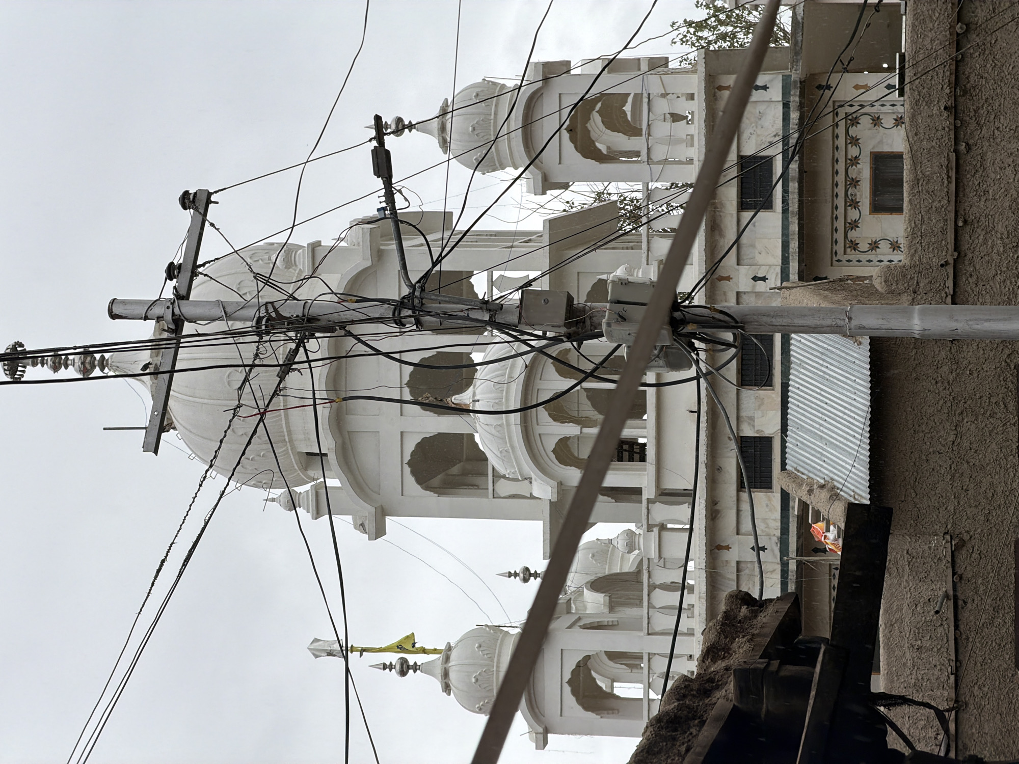 The image depicts the exterior of a white Sikh Gurdwara in Leh, India, against an overcast sky. Prominently in the foreground, a complex network of electrical wires and a utility pole obscures much of the building. The Gurdwara features multiple domes, arches, and decorative finials. A yellow flag flies from one of the pinnacles. Below the Gurdwara, a brown, textured wall and a corrugated metal roof are visible. The overall scene conveys a sense of urban density with interwoven infrastructure and architecture.