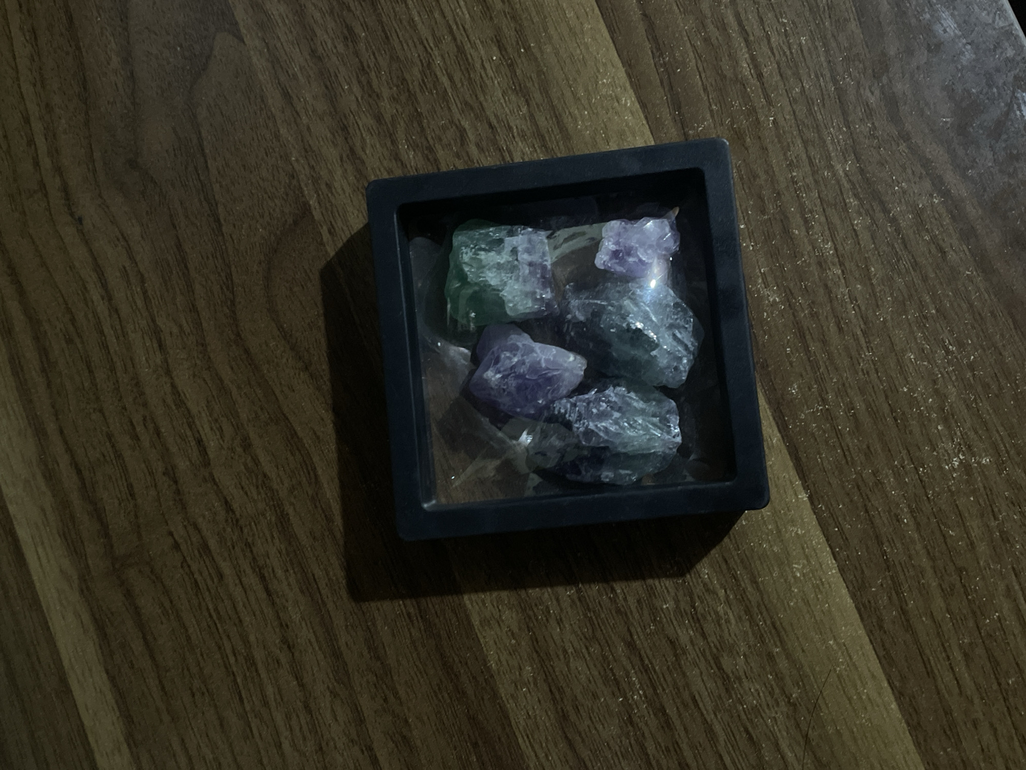 The image captures a collection of raw, unpolished crystals contained within a small, square, black tray. The crystals display a range of colors, predominantly shades of green and purple, along with some clearer or lighter blue pieces. They have an irregular, fractured, and rough texture.
The black tray rests on a dark brown wooden surface, which exhibits a noticeable grain pattern running diagonally across the frame. The lighting in the scene is dim, casting a distinct shadow from the tray onto the wooden surface, indicating a light source from the upper left. No people are visible, and no specific activity is taking place; it is a still life composition. There is no visible text in the image.