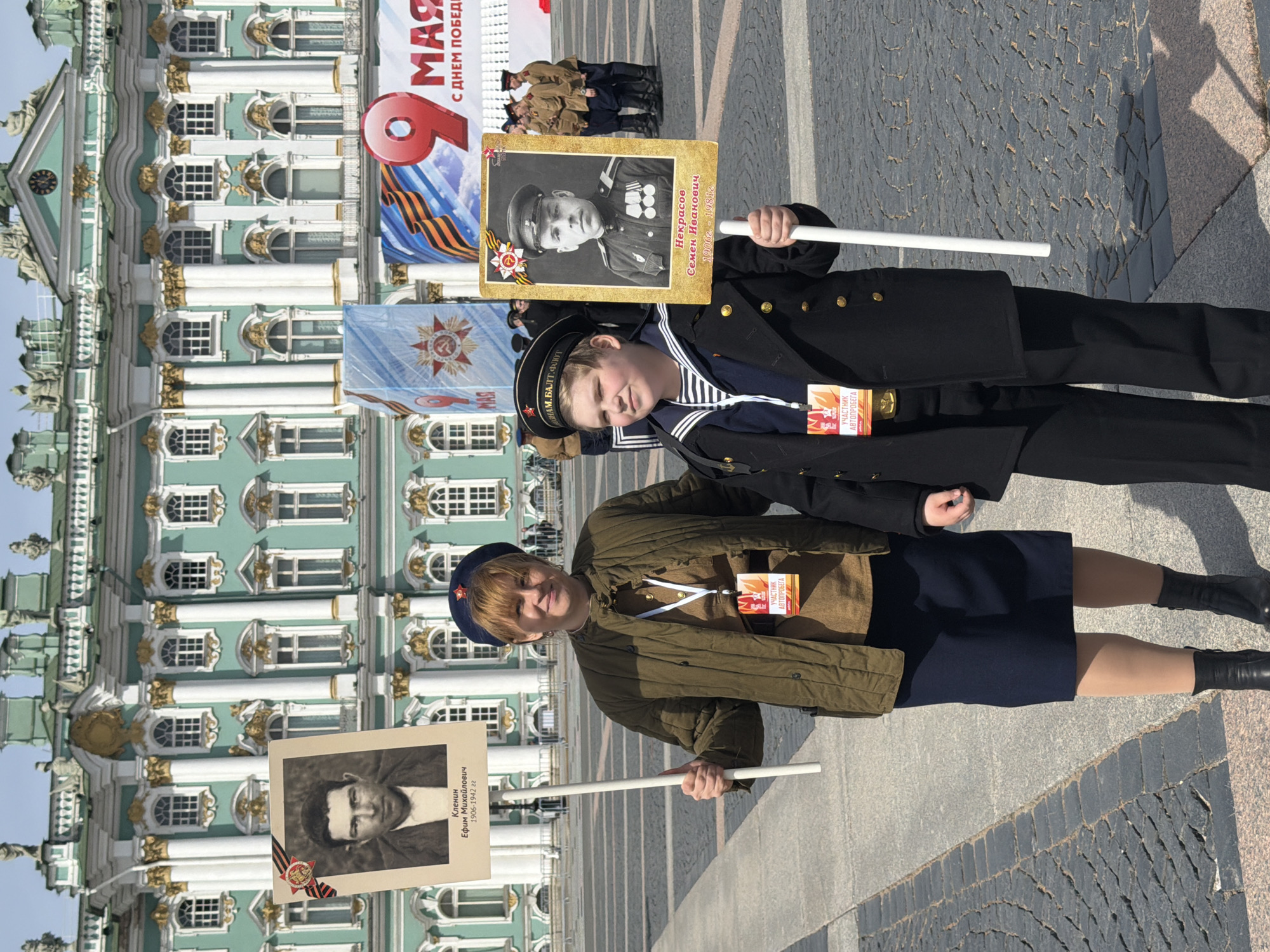 In the city of Saint Petersburg, Russia, two individuals stand in what appears to be a commemorative procession, likely related to Victory Day. A woman on the left, with short blonde hair and a blue beret, is dressed in military-style attire, holding a sign bearing the portrait of a man identified as "Кленин Ефим Михайлович 1906-1942 гг." A younger boy, dressed in a naval uniform, stands on the right holding a similar sign with the portrait of "Некрасов Семен Иванович 1906г. - 1981г." Both are wearing tags that say "Участник Автопробега." Behind them is a building that appears to be the Hermitage Museum and a Victory Day banner.