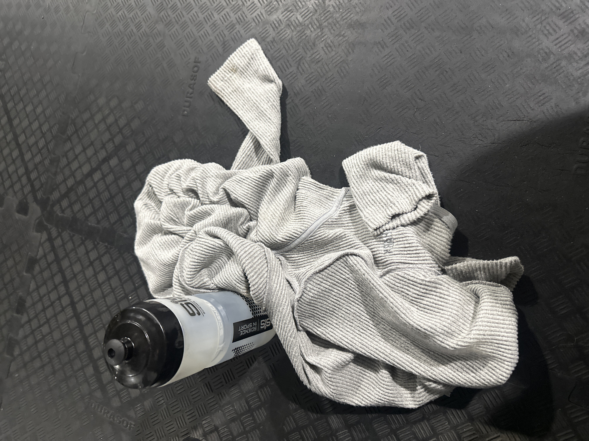 The image depicts a black water bottle with a black nozzle lying on a black, textured rubber floor. The bottle has the words "SiS SCIENCE IN SPORT" printed on it in white. A light gray, folded sweatshirt with a zipper is crumpled next to and partially on top of the bottle. The background consists of the same black, textured rubber flooring. The location is potentially within Tonbridge and Malling, United Kingdom.