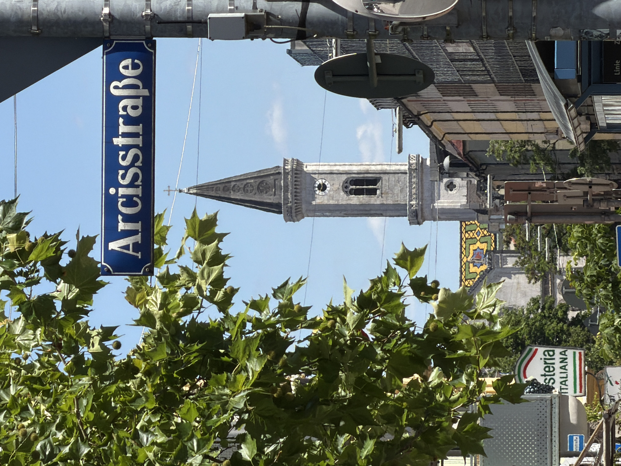 In Munich, Germany, a street sign reading "Arcisstraße" is visible against a bright blue sky. The sign is mounted on a metal pole, and a portion of a large, leafy tree with seed pods partially obscures the view. In the background, a tall, gray church tower with a clock face and a cross on top stands prominently. Adjacent to the tower are buildings with various architectural styles and window patterns. A sign for "osteria ITALIANA" is also visible, indicating a nearby restaurant. The scene depicts a typical urban streetscape with a blend of historical and modern elements.