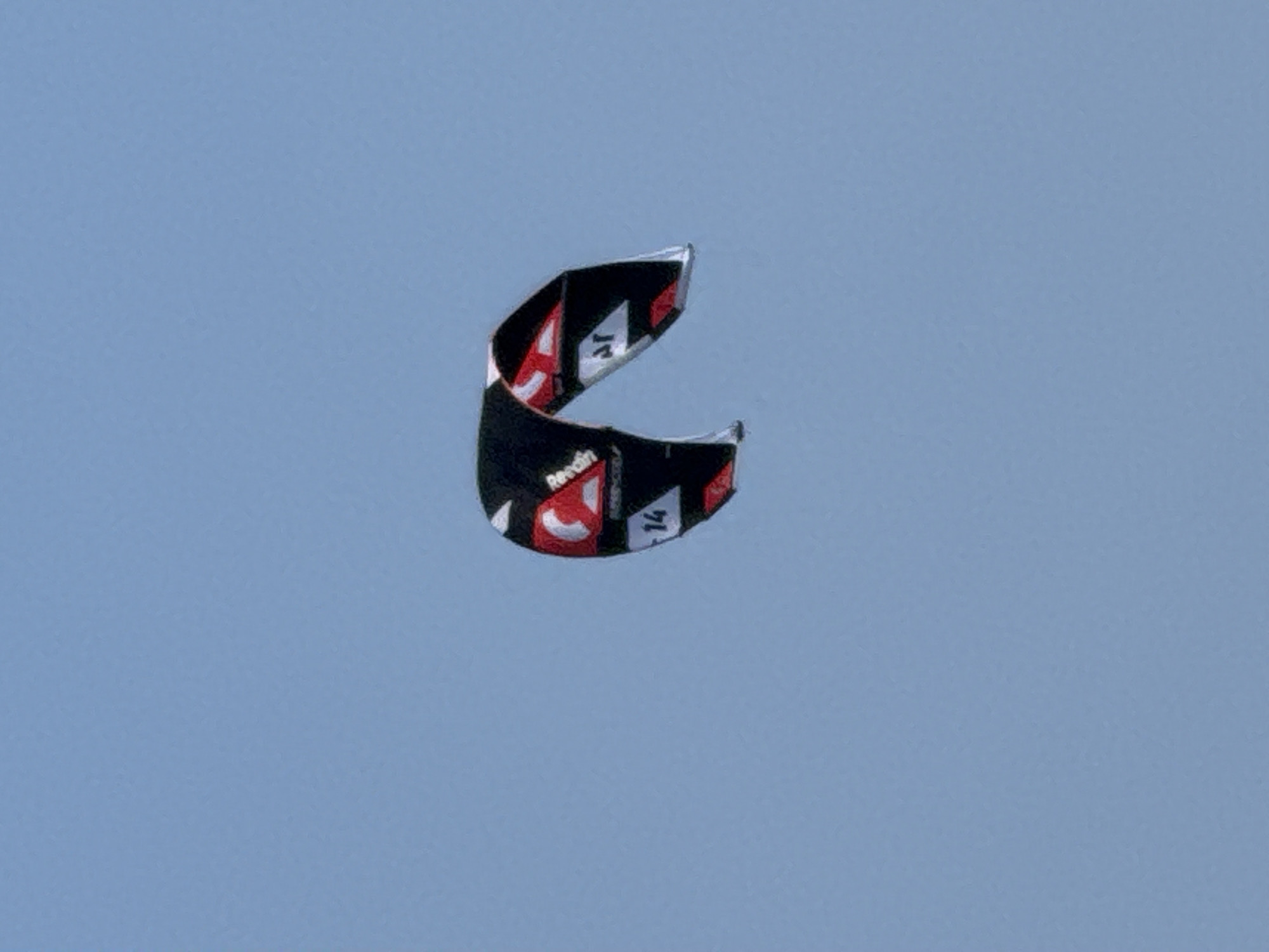 A black and red kite is suspended in a clear, light blue sky. The kite is curved, with a portion of its underside visible. It features white lettering and red accents. A portion of the number "14" is visible in white on the black surface of the kite. The kite is positioned in the center-left of the frame against a uniform sky. The scene is devoid of any other objects, structures, or individuals. The image appears to have been taken from a low angle, looking up at the sky. There is no discernible information within the image that would indicate its specific location within Denpasar, Indonesia, or any other urban environment.