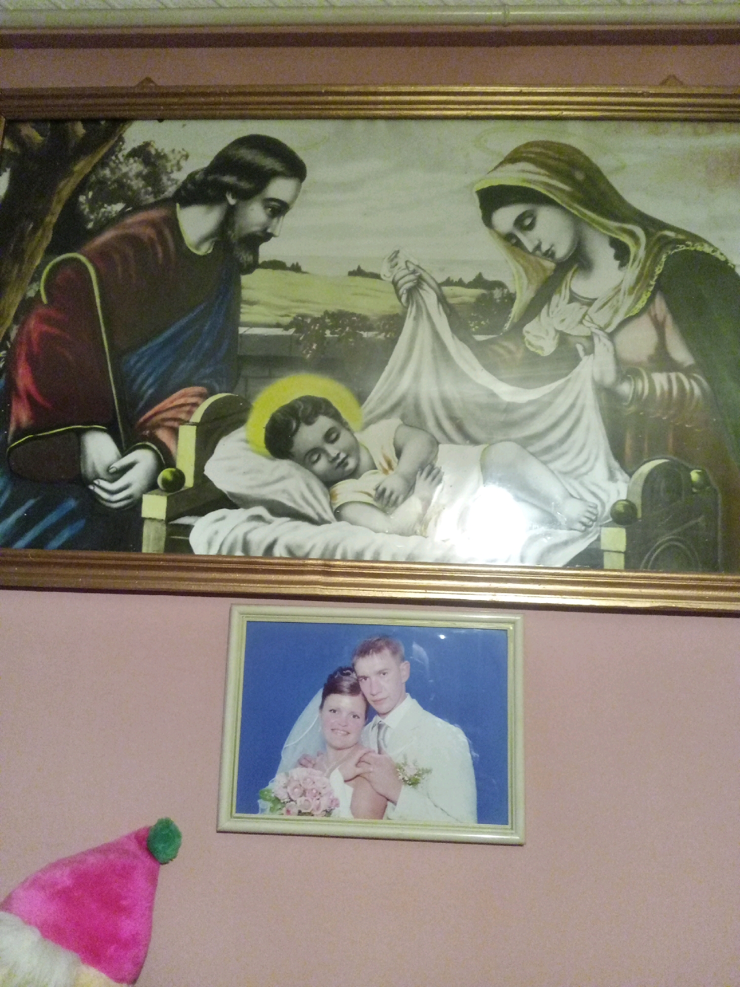 The image shows two framed pictures hanging on a pink wall. The upper frame contains a religious painting depicting the Holy Family. Jesus, as a baby, is lying in a manger, with Mary and Joseph looking over him. The lower frame contains a wedding photograph of a couple. The man is wearing a white suit, and the woman is wearing a wedding dress and veil, holding a bouquet of pink flowers. To the left of the frame, a pink Santa hat is visible. The overall impression is that these are decorative items in a home environment. There is no specific indication of location in the city of Tiachiv, Ukraine.