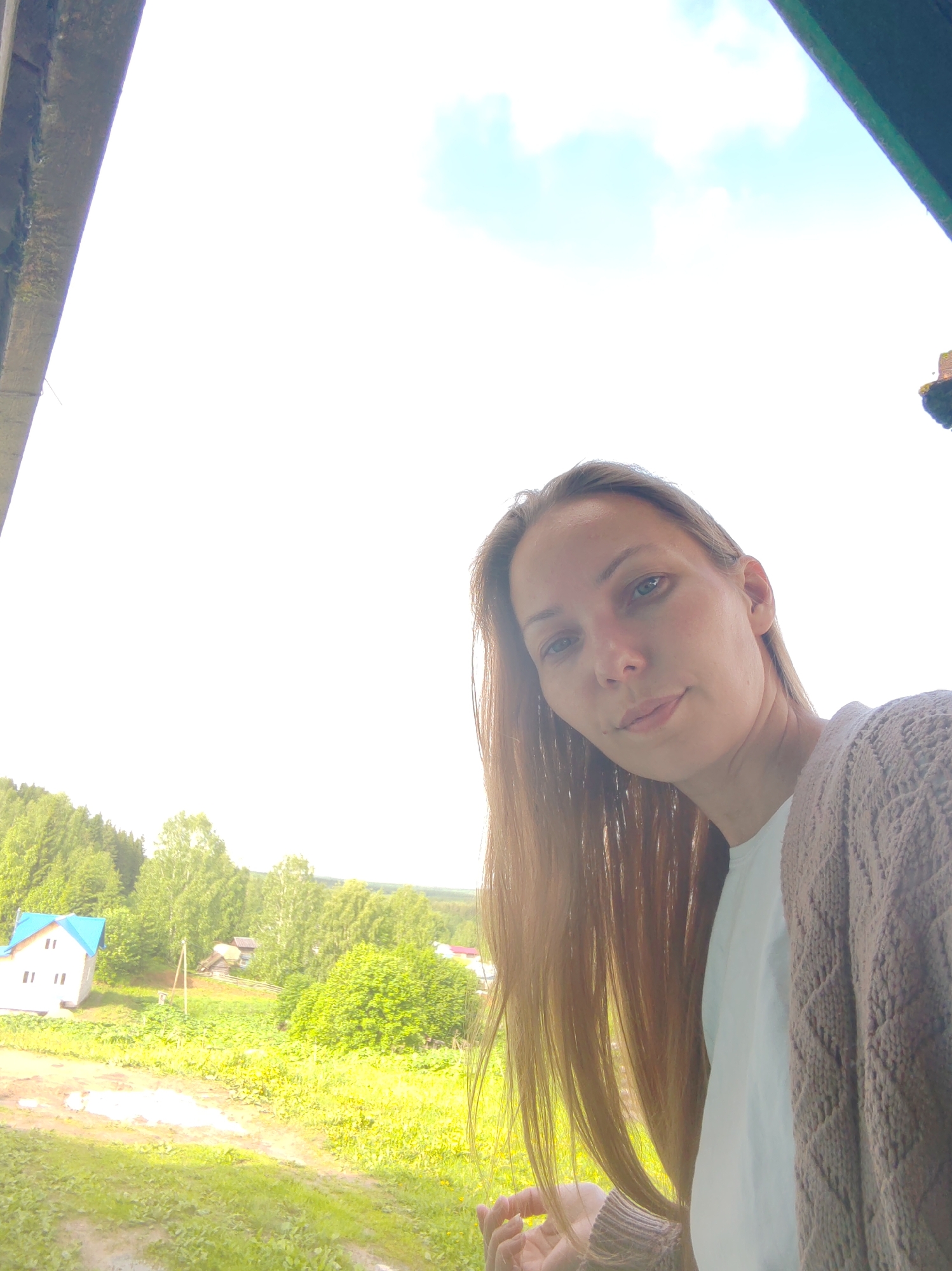A woman with long, straight blonde hair is the subject of the photograph. She has fair skin, blue eyes, and a neutral expression. She is wearing a white t-shirt and a brown cardigan.
The background of the image features a rural landscape, including a bright green field and various trees, some of which appear to be deciduous and others coniferous. A white house with a blue roof is visible on the left side of the frame.
The photo appears to have been taken from an elevated position, as part of a frame of a building is visible at the top of the image. The location is the city of Чухлэм, Russia.