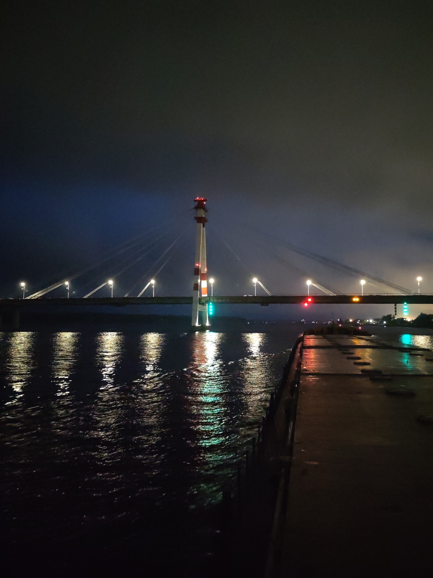 The image shows a bridge at night, with lights reflecting off the water. The bridge has a distinctive tower-like structure in the middle, striped with red and white. Cables extend from the tower, supporting the bridge deck. There are other lights along the bridge, some red, yellow, and green. The water is dark and choppy, reflecting the lights from the bridge and nearby structures. In the foreground, there appears to be a pier or dock, dark and wet. It is unknown what city and country this is taken in.