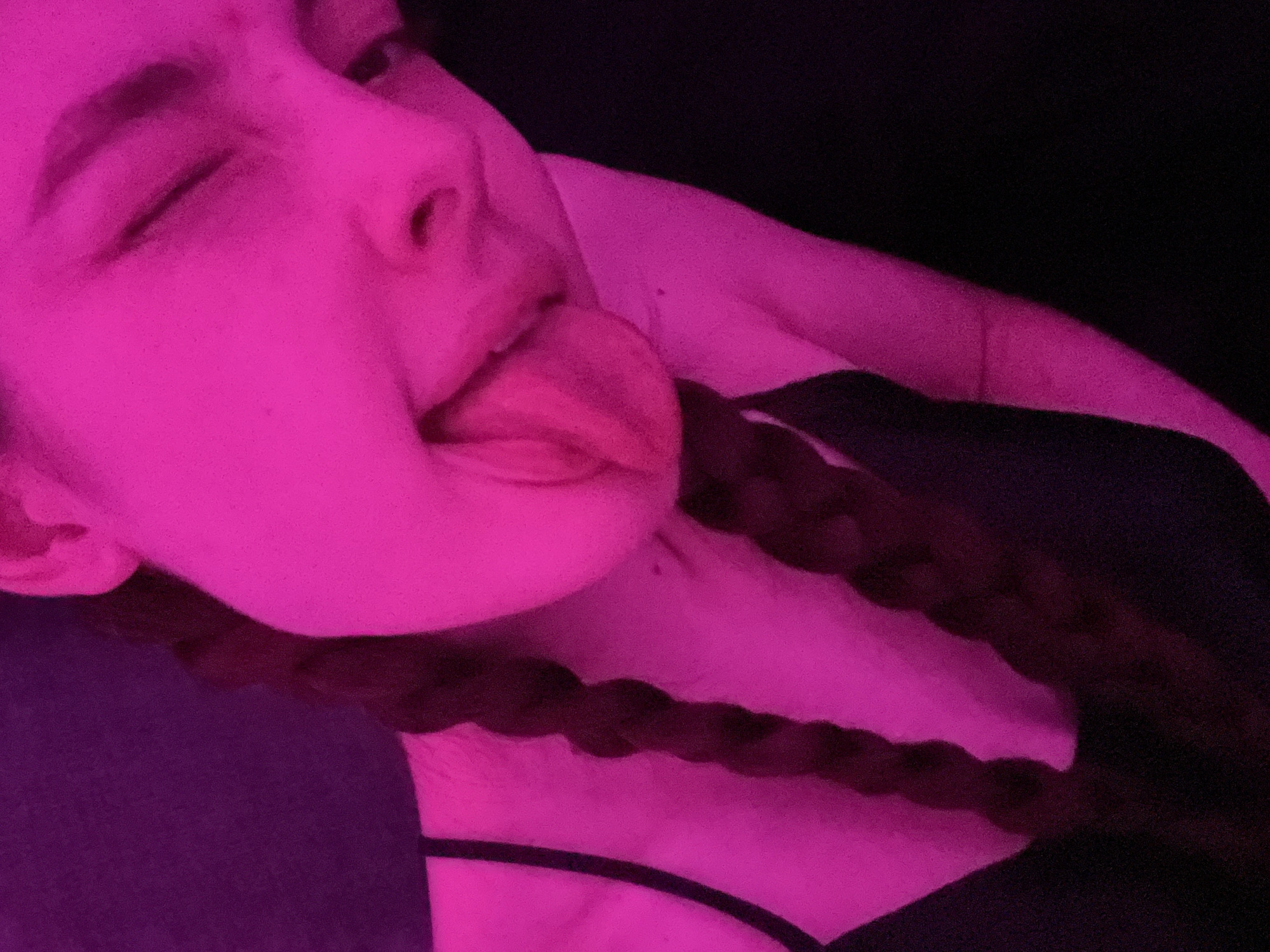 A person is illuminated in pink light, with their tongue sticking out. They have dark hair styled in a braid that is draped across their neck and upper chest. The person is wearing a black camisole. The location is not discernible from the image.