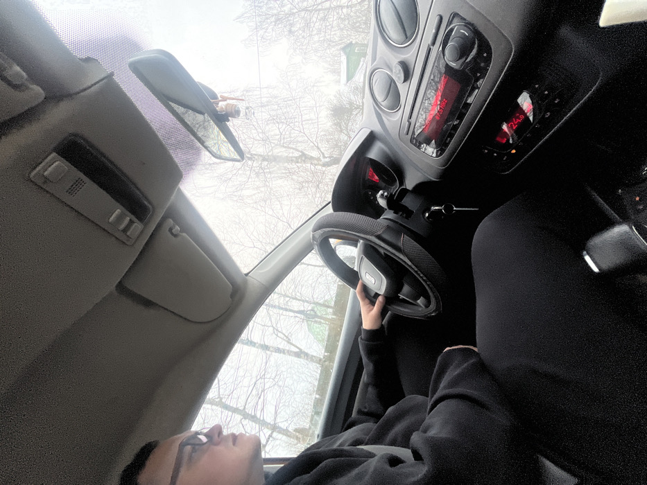 The image presents an interior view from the front passenger seat of a vehicle, looking towards the driver and through the front windshield. A male driver, visible from the mid-torso up, is positioned in the left foreground, wearing glasses and a dark hooded garment. His left hand is resting on the steering wheel, which is covered with a dark, textured material and displays a SEAT logo. Keys are visible hanging from the ignition. The dashboard features a car radio displaying "94.5 MHZ" and "BASTILL," below which a climate control panel shows "24.0." Through the windshield, a cold, overcast outdoor environment is visible, characterized by numerous bare deciduous trees and hints of distant structures, including a building with a green roof, against a hazy sky. This scene is situated in Belavino, Russia.