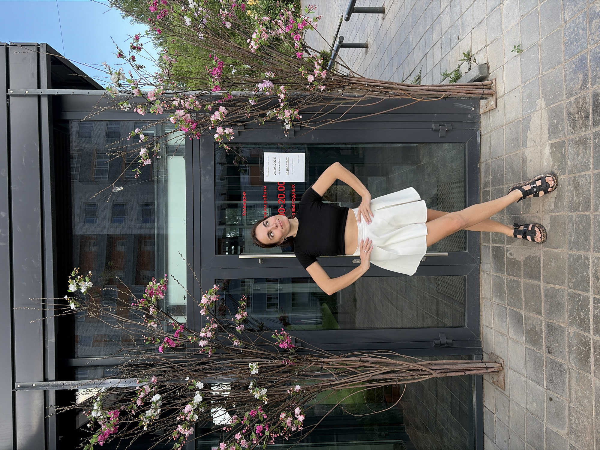 A woman stands in front of a glass-paneled door adorned with floral arrangements. She wears a black short-sleeved shirt and a white skirt, and black sandals. She is positioned with hands on her hips, looking towards the upper-left. The door is flanked by two branches with pink and white flowers, set in the pavement. The doorway appears to be the entrance to a building reflecting a multi-story residential structure. A sign is visible on the door in Russian. The setting is Izhevsk, Russia. The ground is paved with square tiles.