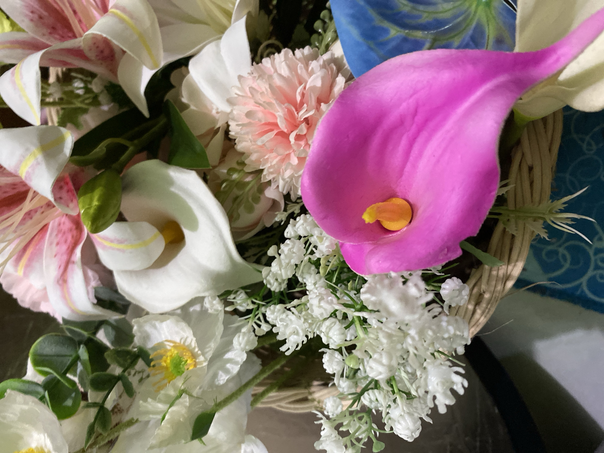 The image shows a close-up of a floral arrangement in a woven basket. The arrangement features several types of artificial flowers, including white calla lilies, pink and white lilies with pink streaks, small white filler flowers resembling baby's breath, and a pink carnation-like flower. A prominent bright pink calla lily with a yellow center is in the foreground, partially obscuring other flowers. The basket is woven from light-colored material and sits on a surface with a blue, patterned object visible in the background. The lighting suggests an indoor setting. The image does not contain any information or context related to Kuala Pembuang, Indonesia.