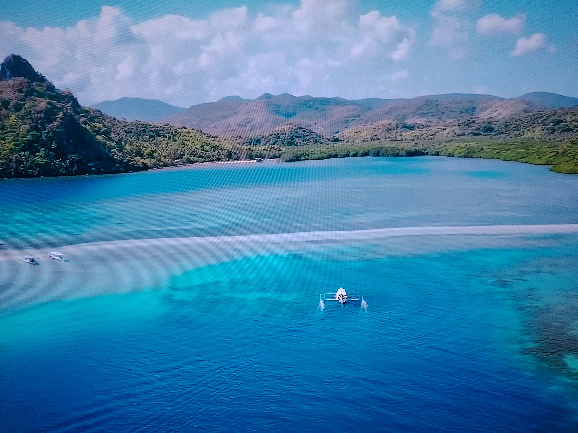 An aerial, high-angle drone shot captures a tranquil tropical scene. A body of bright blue, turquoise, and deep blue water dominates the frame, with a pale sandbar visible in the middle ground, separating shallow, clear water from the deeper ocean. On the sandbar, two small, white boats are moored. In the foreground, a single, white outrigger canoe with a small canopy and two people on board navigates the deep blue water. In the background, lush green, tree-covered mountains rise, with more distant, hazy mountains forming the horizon under a pale blue sky with scattered white clouds. The location is a tropical island, and there is no connection to Huntingdonshire, United Kingdom.
