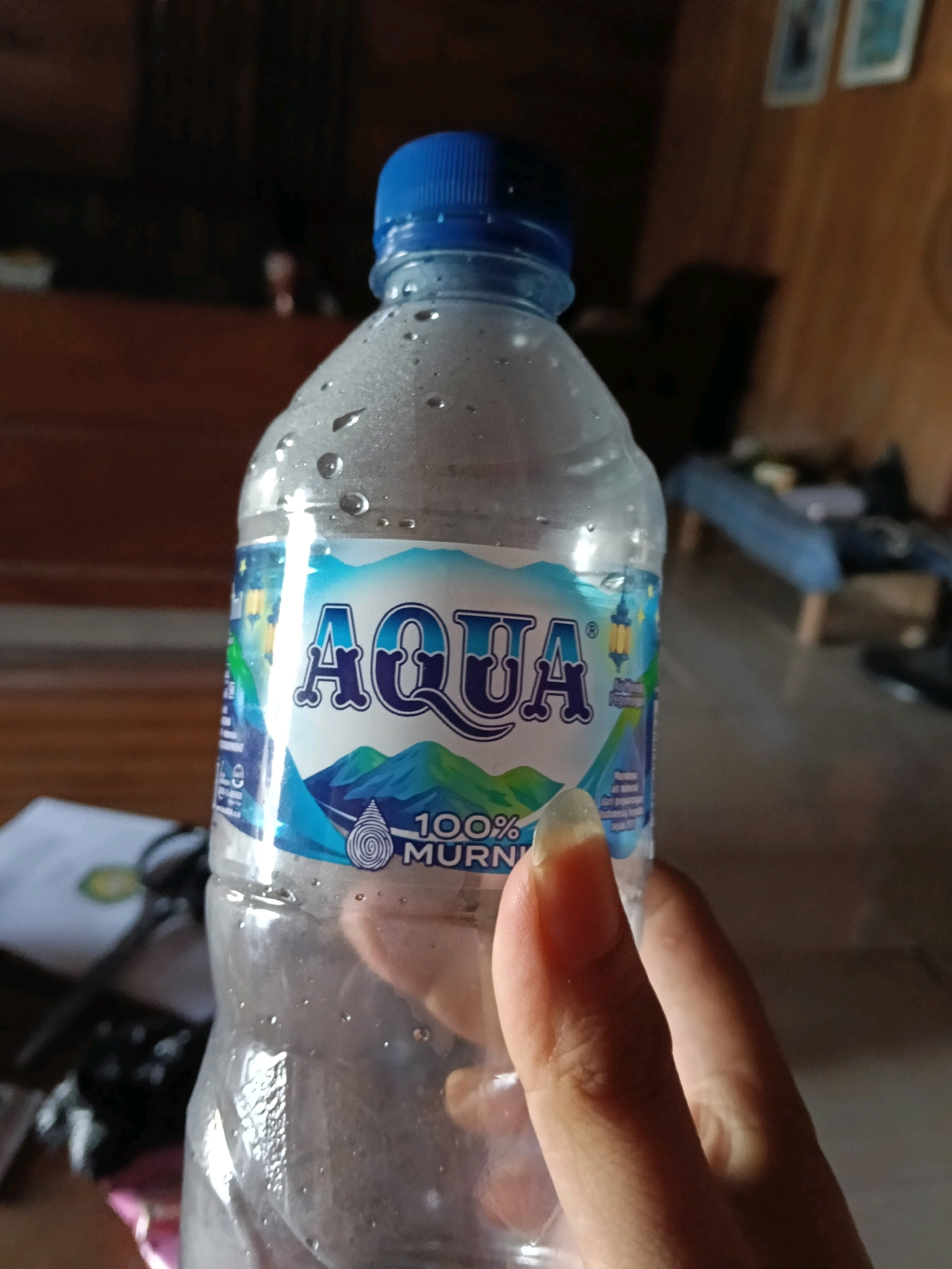 The image is a close-up shot of a person holding a partially empty bottle of Aqua mineral water. The bottle has a blue cap and a label featuring the Aqua logo, mountains, and text that reads "100% MURNI". The bottle is covered in condensation droplets. The background is blurred, showing a wooden surface, some out-of-focus furniture, and what appears to be a wall with some framed pictures. The lighting suggests an indoor setting, possibly a home. The location in Galuk, Indonesia, is not directly depicted or inferable from the visual information present in the image itself.
