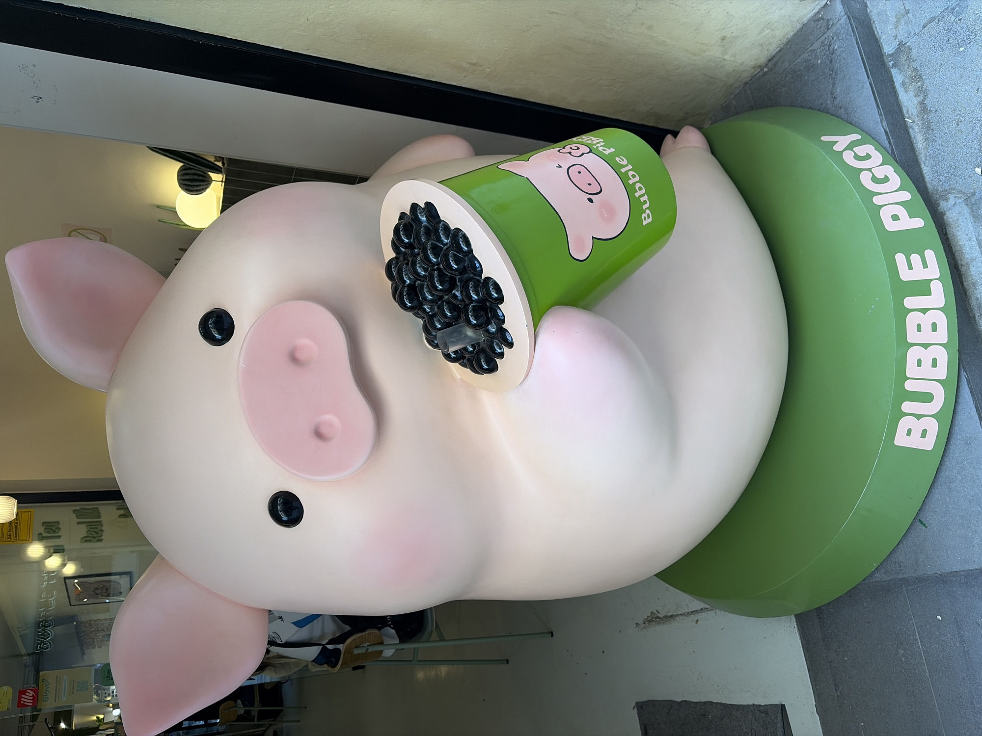 The image depicts a large, anthropomorphic pig statue advertising a "Bubble Piggy" bubble tea establishment. The pig is pink with large, floppy ears and a rounded body. It is holding a green cup filled with black tapioca pearls, designed to resemble a bubble tea drink. The base of the statue is a large, green, cylindrical shape labeled "Bubble Piggy." The statue appears to be located outside the bubble tea shop, evidenced by the storefront visible in the background. The interior of the shop shows signs of tables and chairs, suggesting it is a place where customers can sit and enjoy their drinks. The overall scene suggests a playful and inviting atmosphere intended to attract customers to the bubble tea shop. I am unable to confirm that this is the city of Barcelona, Spain based on the information provided.