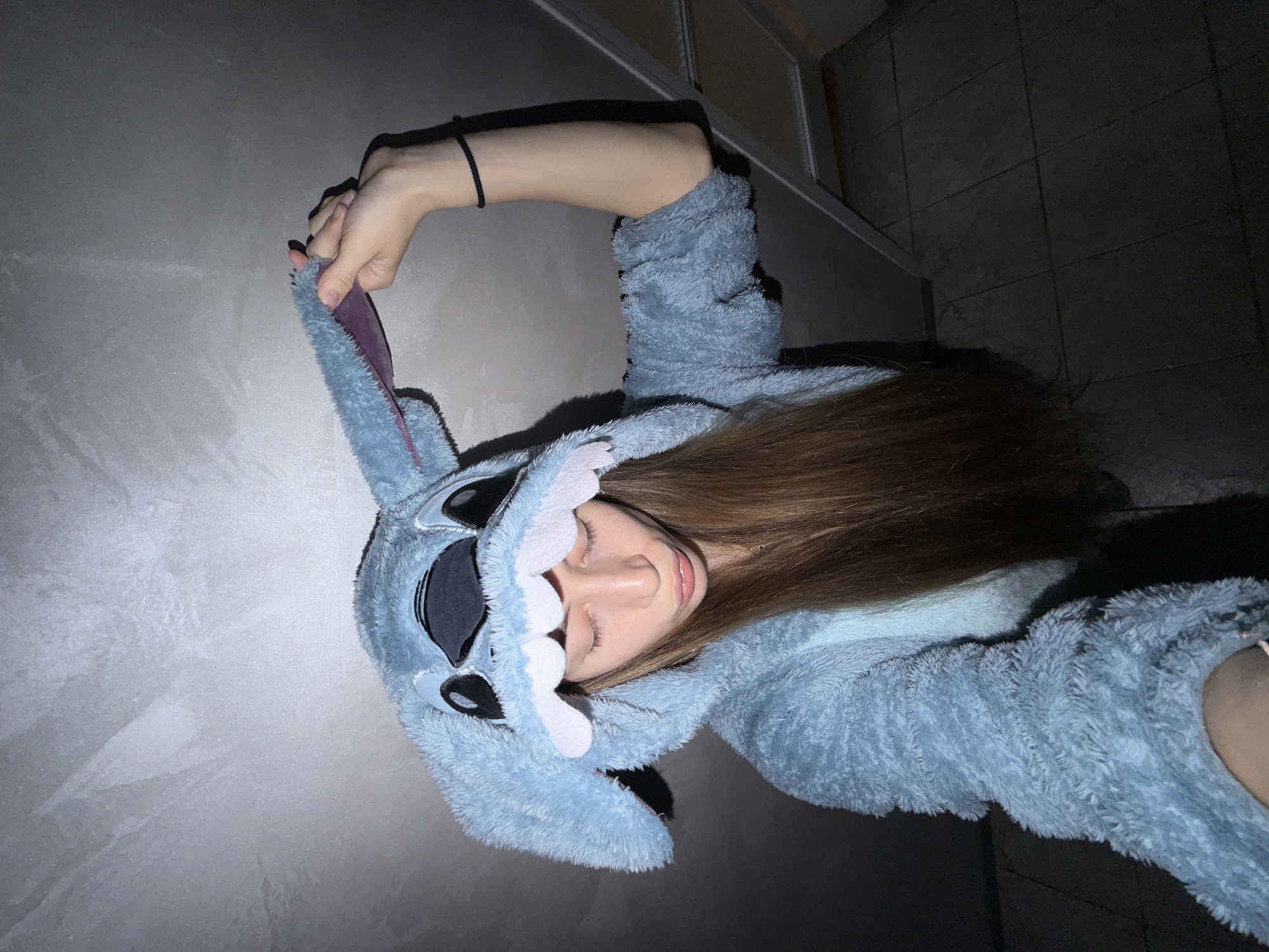 A person wearing a blue, furry Stitch onesie lies on a light gray surface. The person's face, with closed eyes and brown hair, is visible within the onesie's hood. The person's left hand holds the Stitch ear. The background reveals tiled flooring near a wall or edge. The location is not discernible as Plovdiv, Bulgaria, from the visible details.