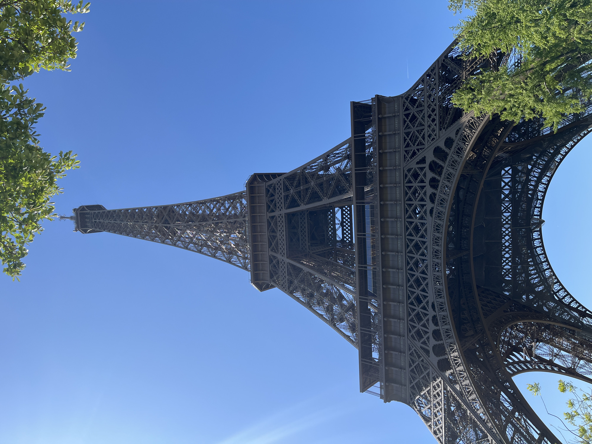 An upward-looking photograph captures the majestic Eiffel Tower in Paris, France, from a low, worm's-eye perspective. The iconic iron lattice structure dominates the frame, tapering towards its peak against a brilliant, clear blue sky. Lush green tree foliage partially frames the tower on both the left and right sides, adding a natural element to the urban landmark.
The intricate details of the tower's ironwork are clearly visible, particularly on the lower and middle sections. Engraved names of French scientists and engineers are discernible along the first level's perimeter. Visible names include "BORDA," "EYCRAT," "FOURIER," "SAVAGE," "PELOUZE," "CARNOT," and "LAMÉ." Other names like "BOUSSINGAULT," "PERRIER," "BRAZIERE," and "MALEUS" can also be seen further along the structure.
The bright blue sky, devoid of clouds, suggests a sunny, clear day. A faint white contrail from a distant aircraft is visible in the upper sky, slightly to the right of the tower's apex. No people or specific activities are visible, focusing solely on the architectural grandeur of the monument.