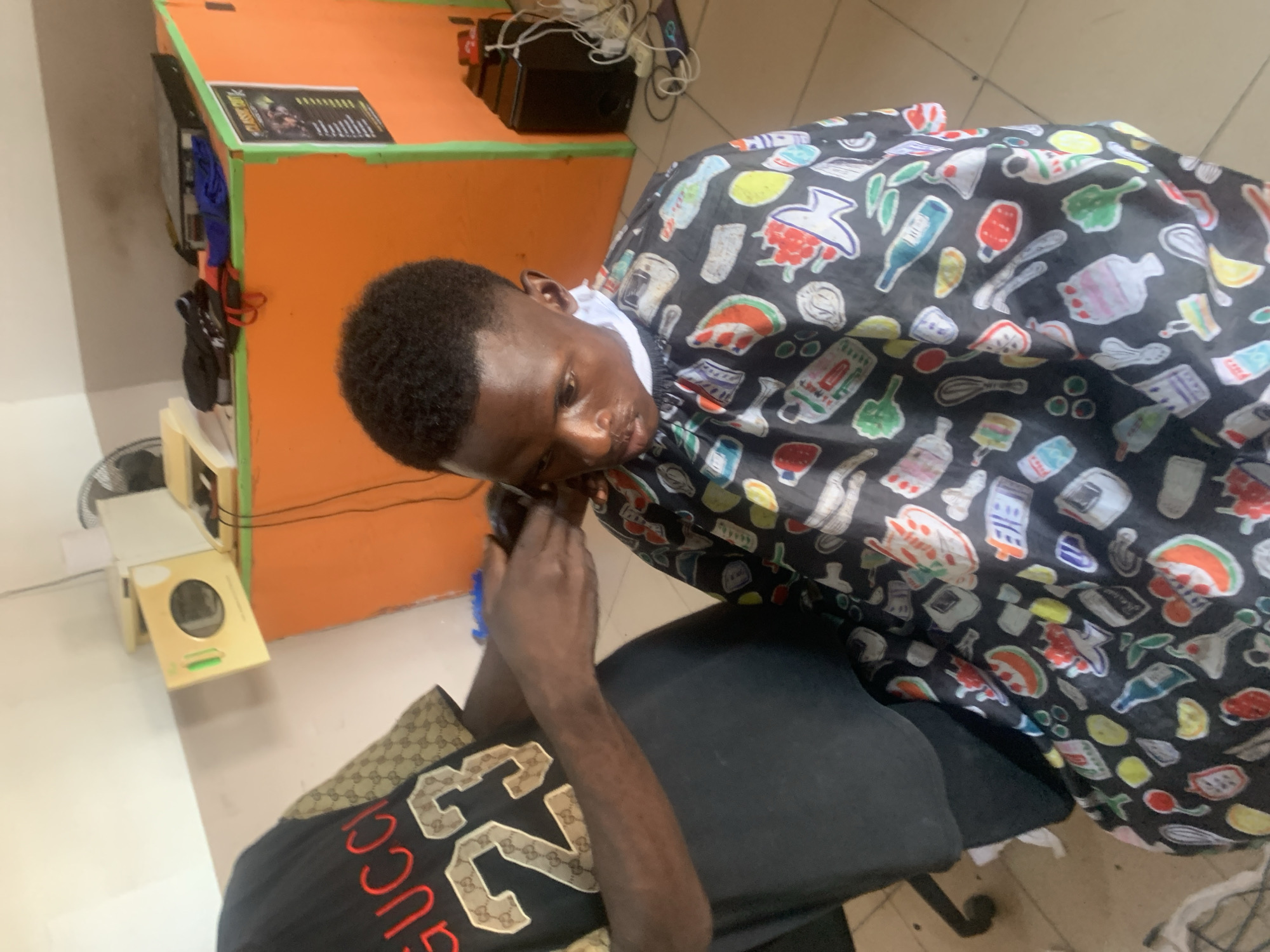 The image shows a young Black man in a barber shop, receiving a haircut. He is seated on a chair, wearing a dark cape with a pattern of various kitchen items, such as bottles, whisk, and food illustrations. The barber, whose hands are visible, is in the process of cutting the man's hair.
The setting appears to be a modest barber shop. In the background, there is an orange counter with a green trim, on which a black speaker and some electrical wires are placed. To the left of the counter, a white and gold equipment is visible. The floor appears to be tiled. The lighting suggests it is daytime, and no information about the weather is available.
The man's expression is neutral as he looks down, likely concentrating on the haircut. His shirt, partially visible at the bottom of the frame, has a brown and gold pattern with red lettering that seems to say "GUCCI".
The image is taken from a slightly elevated angle, looking down at the subjects. There is no discernible text in the image that provides further context about the location or the individuals. The presence of the barber's tools and the haircutting activity clearly indicate a commercial service is being provided.