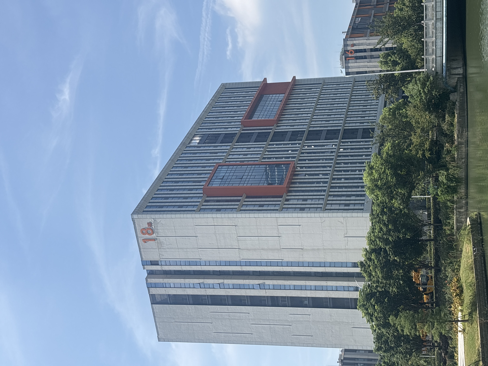 The image displays a modern, multi-story building complex in the city of Suzhou Industrial Park, China, under a clear blue sky with wispy clouds. The main building, a prominent structure, features a light-colored facade with vertical window arrangements and striking orange accents. A large orange numeral "18" is visible on the upper portion of this building, indicating its designation. To the right of this main building, another building with the numeral "16" is partially visible, also with orange architectural elements.
In the foreground, a body of calm water, likely a canal or river, reflects the sky and surrounding greenery. Lush trees and vegetation line the banks of the water and extend up to the base of the buildings. A bridge with decorative railings spans across the water in the mid-ground. A yellow construction vehicle, possibly a crane or excavator, is partially obscured by trees in the lower left portion of the image. The scene suggests a well-developed urban environment with a focus on modern architecture and green spaces. No people are discernibly present in the image.