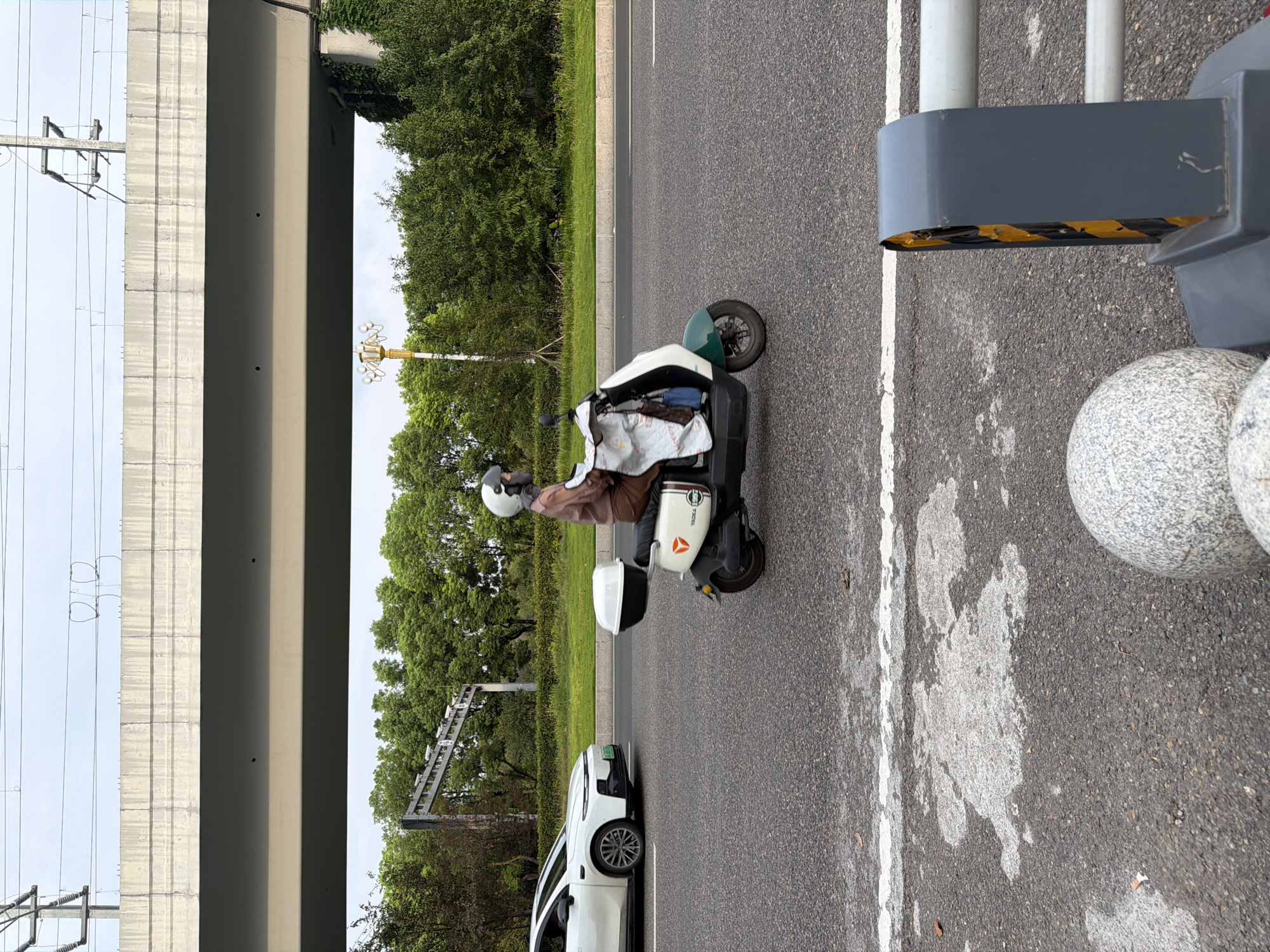 A person wearing a helmet and a brown jacket rides a white and green electric scooter on a paved road. The scooter has a white storage box on the back. The rider is covered by a light-colored blanket draped over them. A white car is visible to the left of the scooter. In the background, there are trees and a concrete overpass with power lines. The scene appears to be an urban setting in Jiangjin, China.