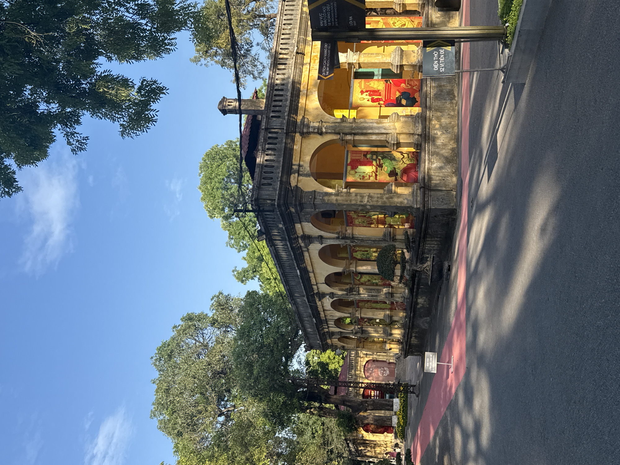The image shows an ornate, yellow building with arched openings in the city of Hà Nội, Vietnam. The building appears to be a historical site, with multiple signs in Vietnamese and English indicating its significance. Some of the signs mention "DI TÍCH CÁCH MẠNG D67 / THE D67 REVOLUTIONARY RELIC" and "ĐIỆN THỜ / 52 VỊ TIÊN ĐỂ." Trees line the street in front of the building. A red line marks the walkway. The sky is visible behind the trees and the top of the building, suggesting it's a sunny day. A narrow asphalt road is visible.