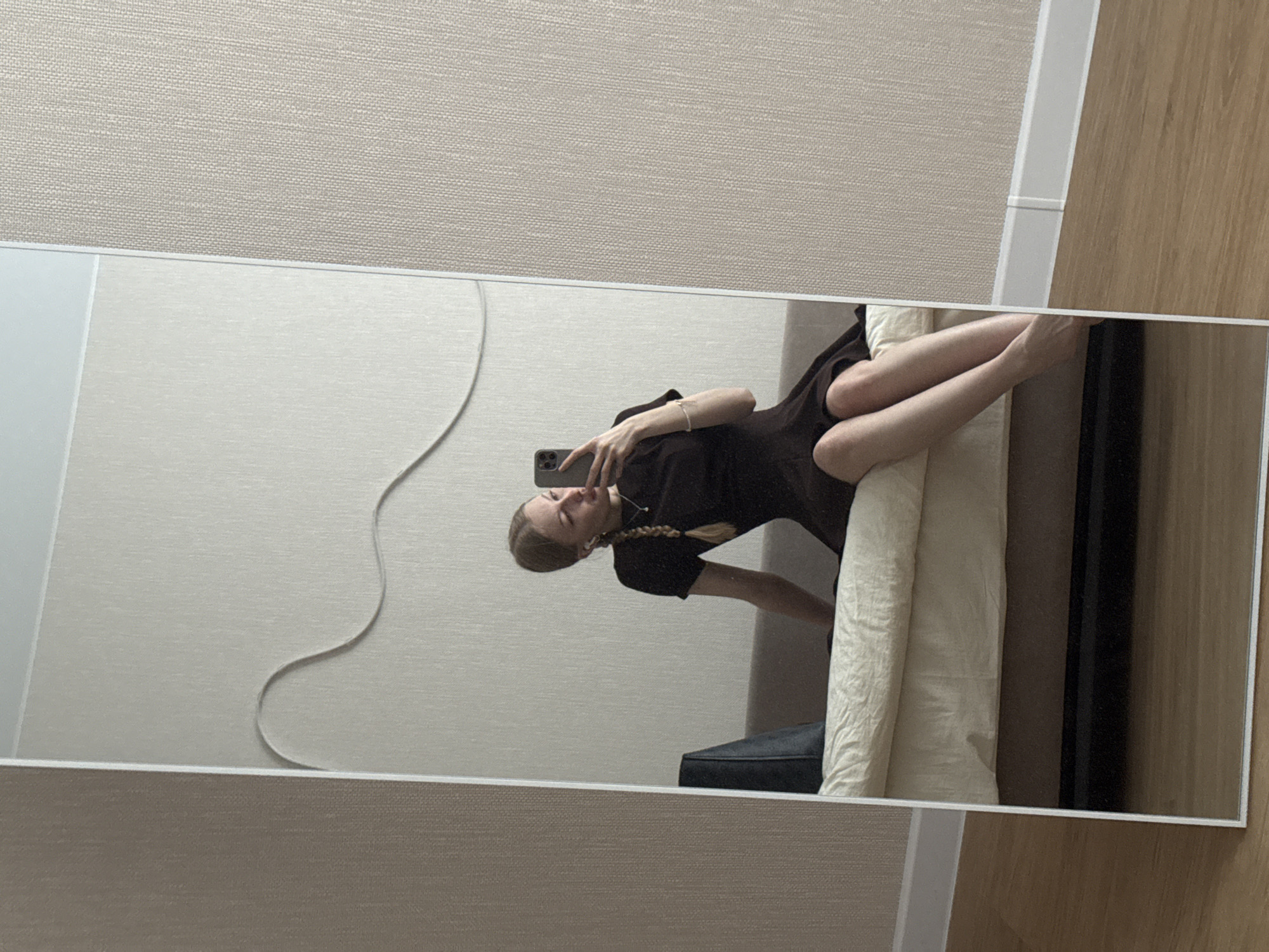 A young woman is reflected in a full-length mirror. She is lying on a light-colored, patterned surface, possibly a bed or couch. She is holding a mobile phone in her right hand, seemingly taking a selfie. She is wearing a dark-colored short-sleeved top and has a braid in her hair. Her left arm is resting on the surface. The background consists of a textured beige wall, white trim, and a portion of a wooden floor. A dark object, possibly luggage, is also visible in the reflection. The location appears to be indoors, possibly a residential space in the city of Perm, Russia.