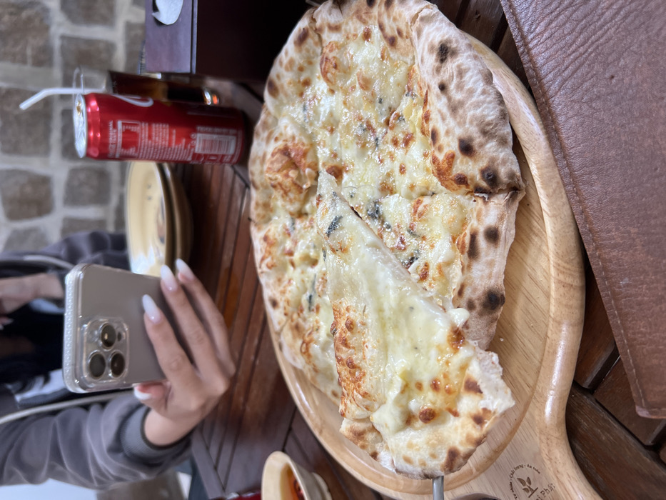 A person with long nails holds a smartphone, photographing a pizza on a wooden paddle. The pizza, featuring a browned crust and a generous amount of melted cheese with blue cheese, has a slice being lifted by a metal server. A can of Coca-Cola with a straw sits nearby, and parts of a yellow plate and sauce dish are also visible on the wooden table.