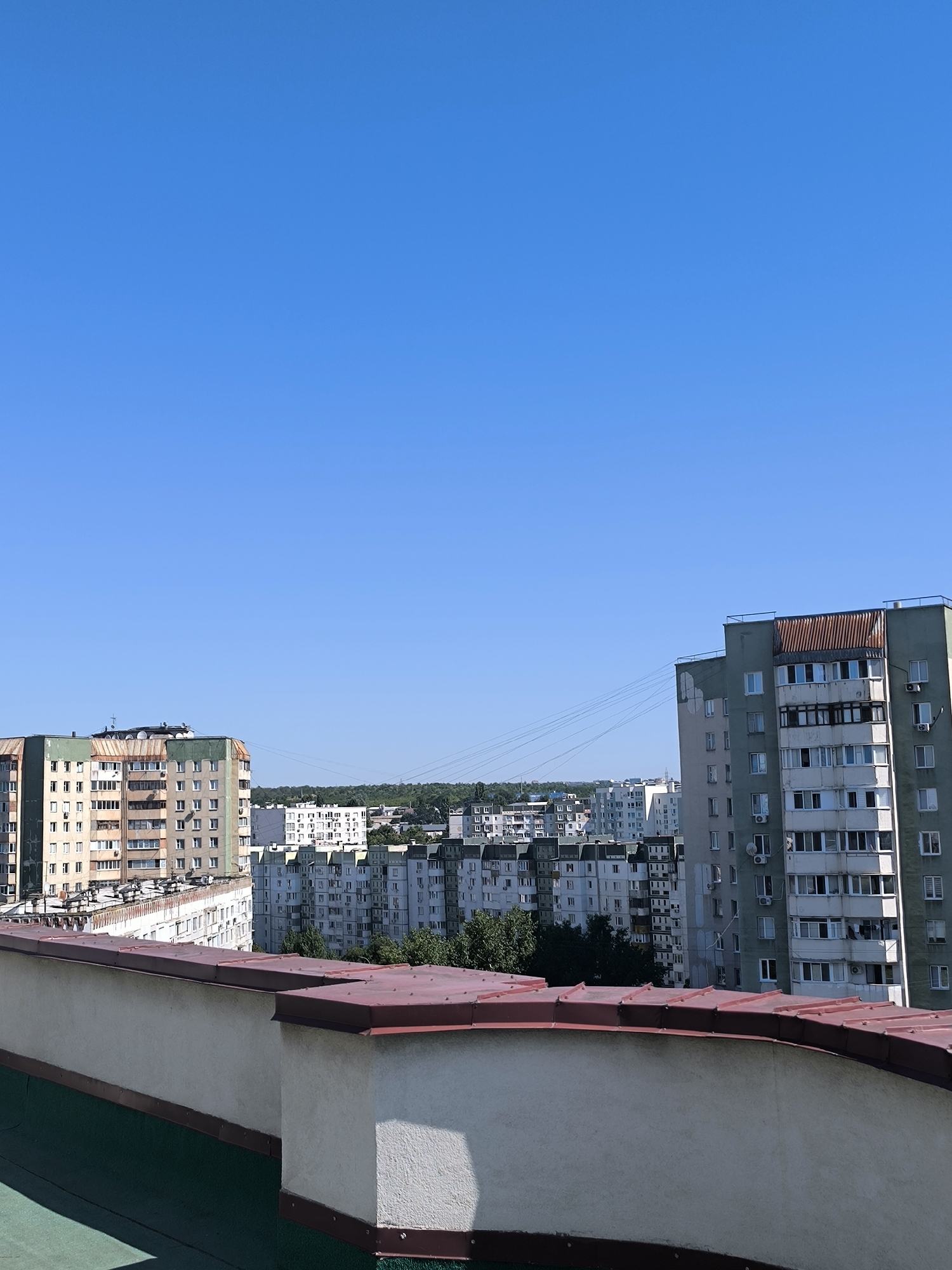 The image depicts a skyline view of apartment buildings in Chișinău, Moldova, under a clear blue sky. In the foreground, a white parapet with a red metal coping runs along the edge of a rooftop. Beyond the parapet, several multi-story apartment buildings are visible. The building on the left is light beige with green accents and appears to be older Soviet-style architecture. To its right, a lighter colored building with numerous windows is partially obscured. Further to the right, a prominent tall, green apartment building with a rusty metal structure on its top floor dominates the middle ground. Behind these buildings, a dense cluster of similar apartment blocks stretches towards the horizon, interspersed with trees and what appear to be power lines crisscrossing the sky. The overall scene suggests an urban residential area on a bright, sunny day.