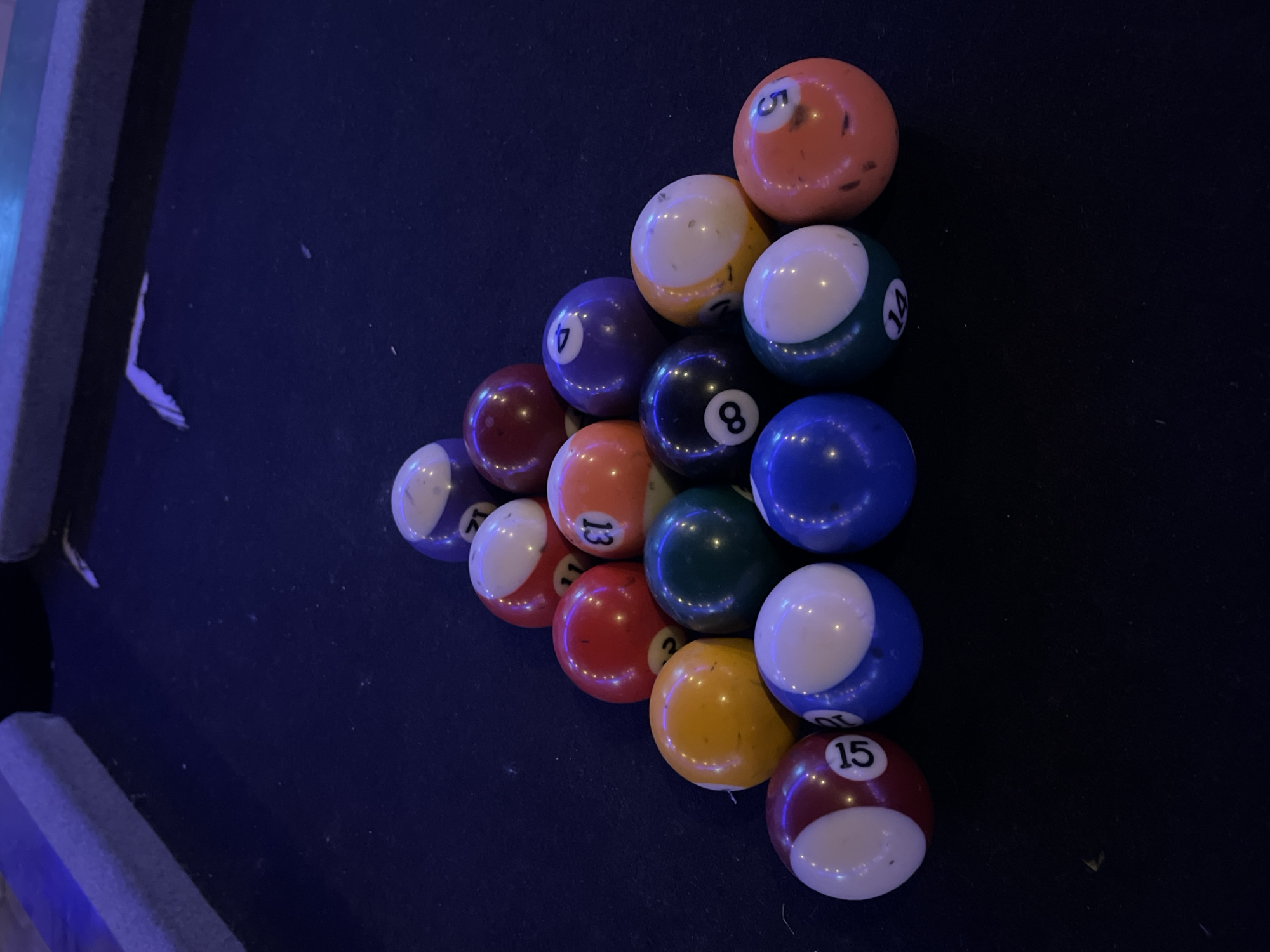 A close-up, high-angle shot depicts a racked set of fifteen billiard balls on a dark blue billiard table. The balls are arranged in a triangular formation. The rail of the pool table is visible on the left side of the frame, indicating an indoor setting. Specific details about people, actions, or the environment beyond the table are not visible. The city and state are not identifiable from the visual information provided.